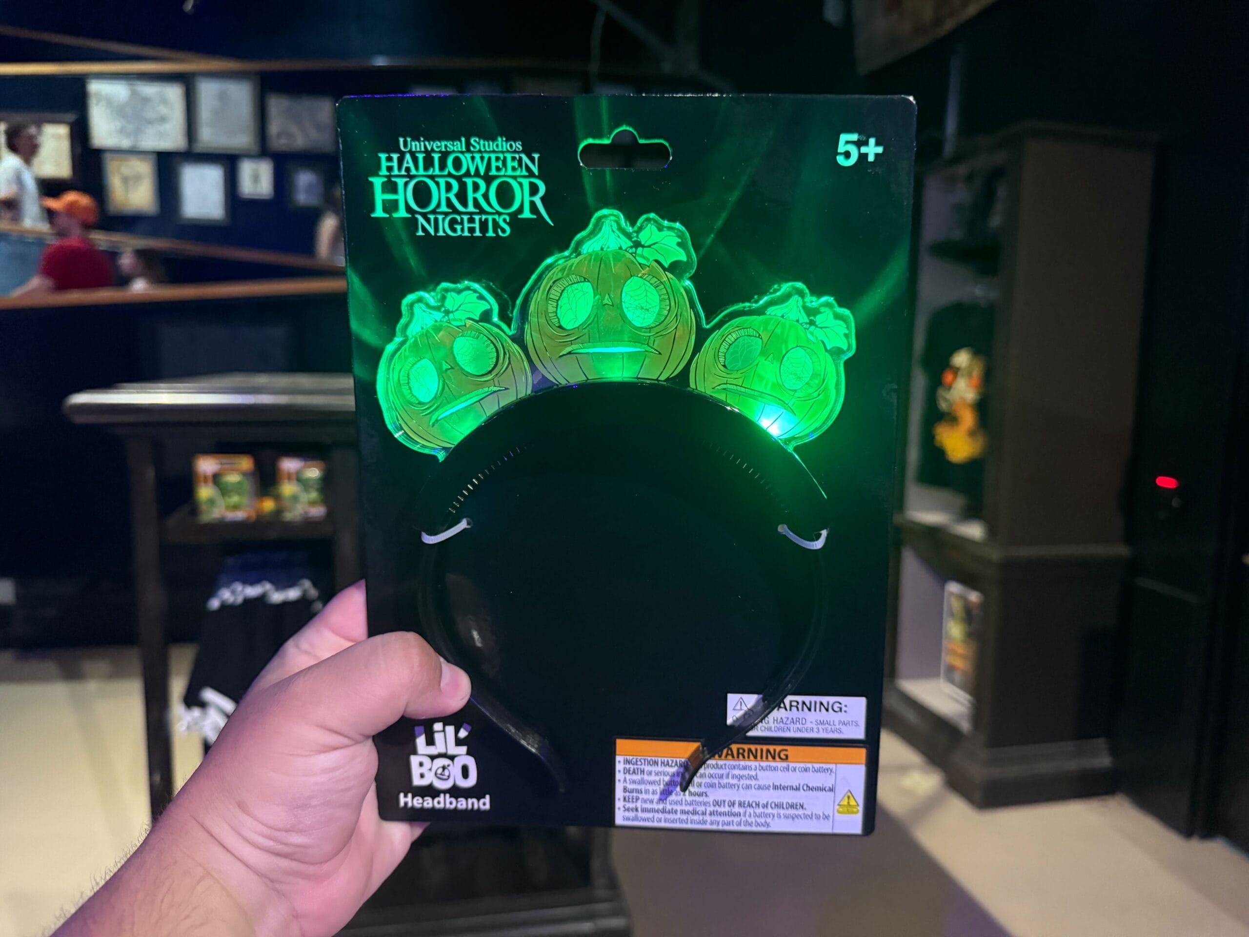 A hand holds a "Lil Boo" pumpkin headband from Universal Studios' Halloween Horror Nights in a store.