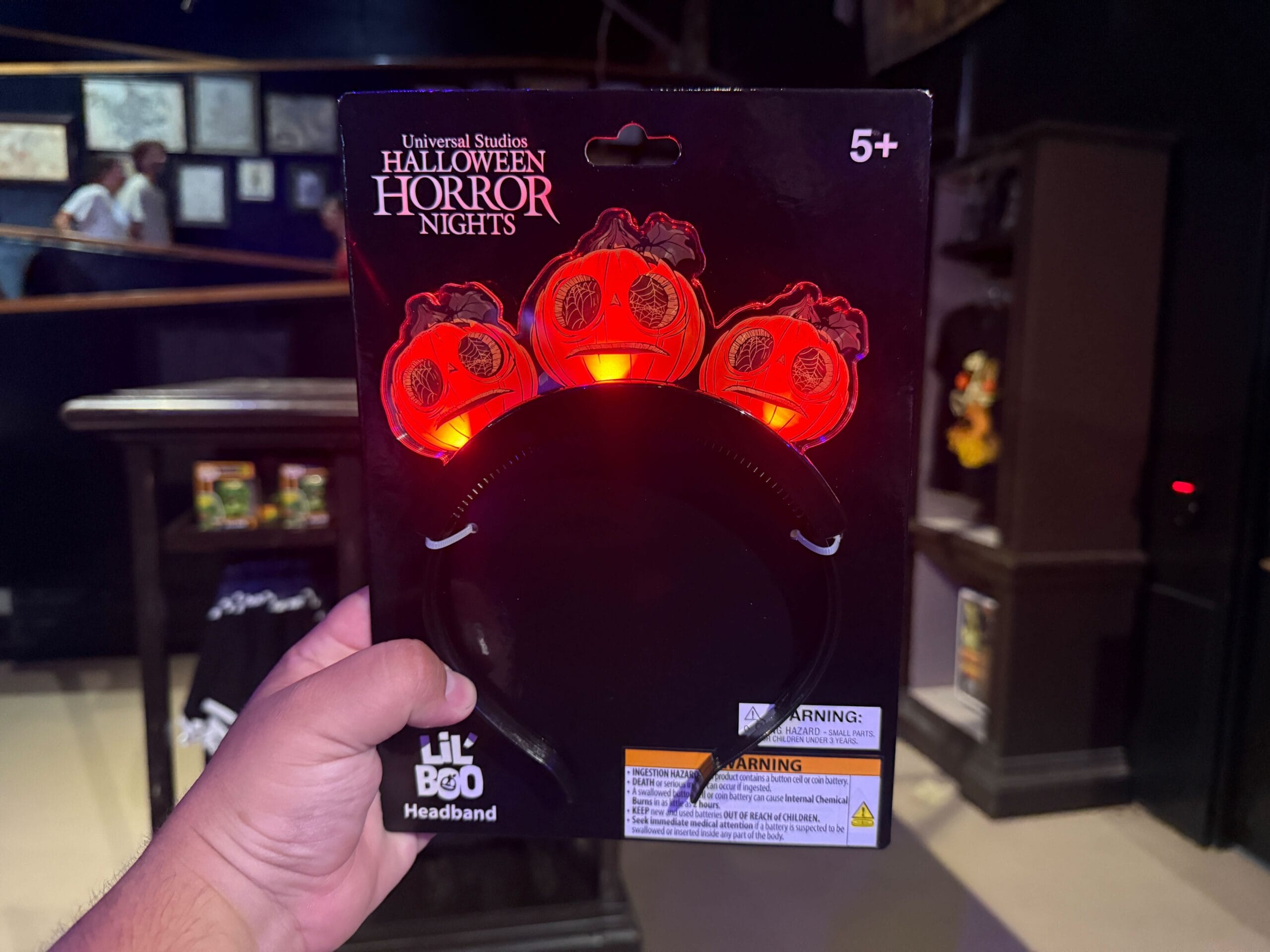 A hand holds a Lil' Boo headband with three glowing jack-o'-lanterns, labeled for Halloween Horror Nights.