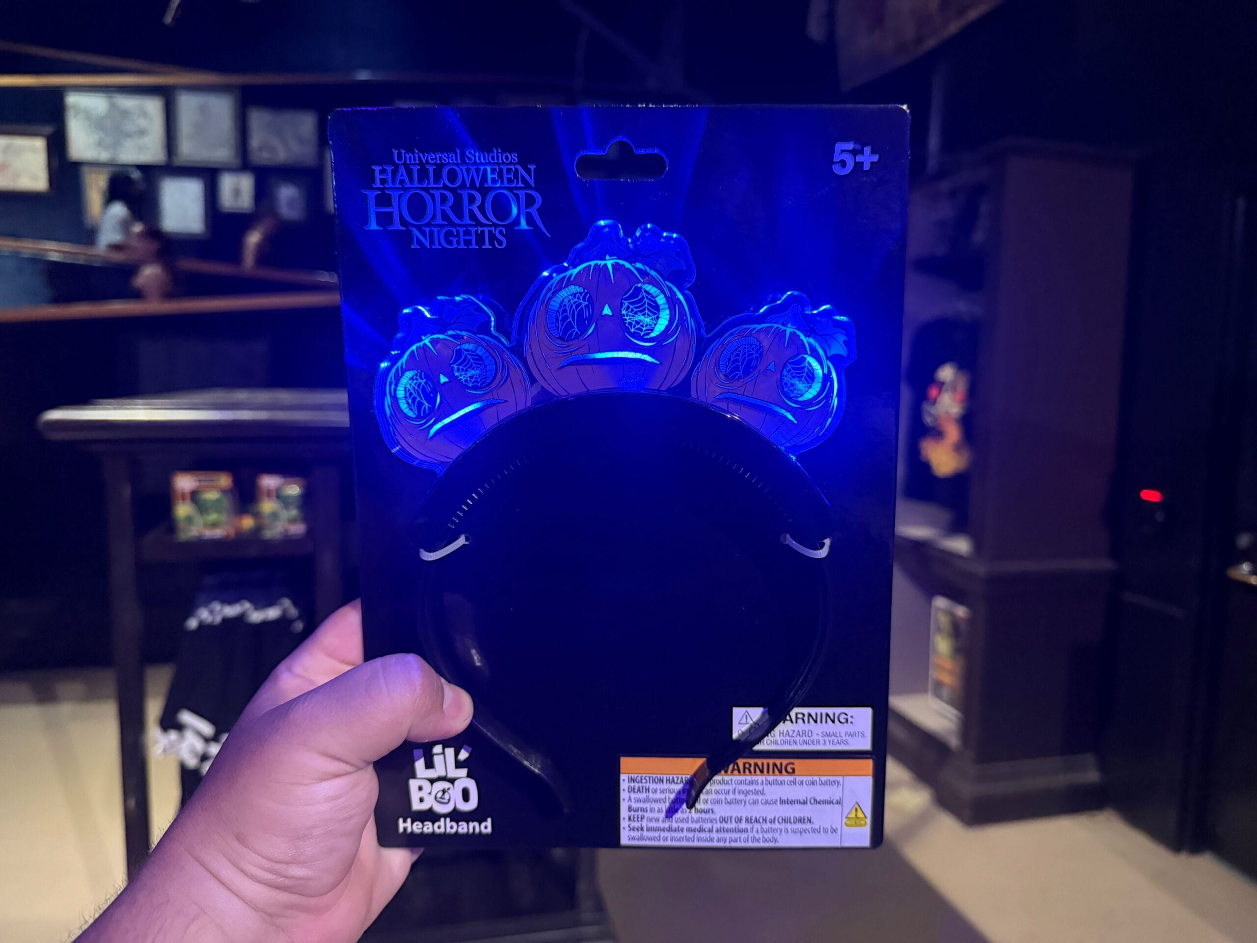 A hand holds a Lil Boo light-up headband package from Universal Studios Halloween Horror Nights in a dimly lit room.