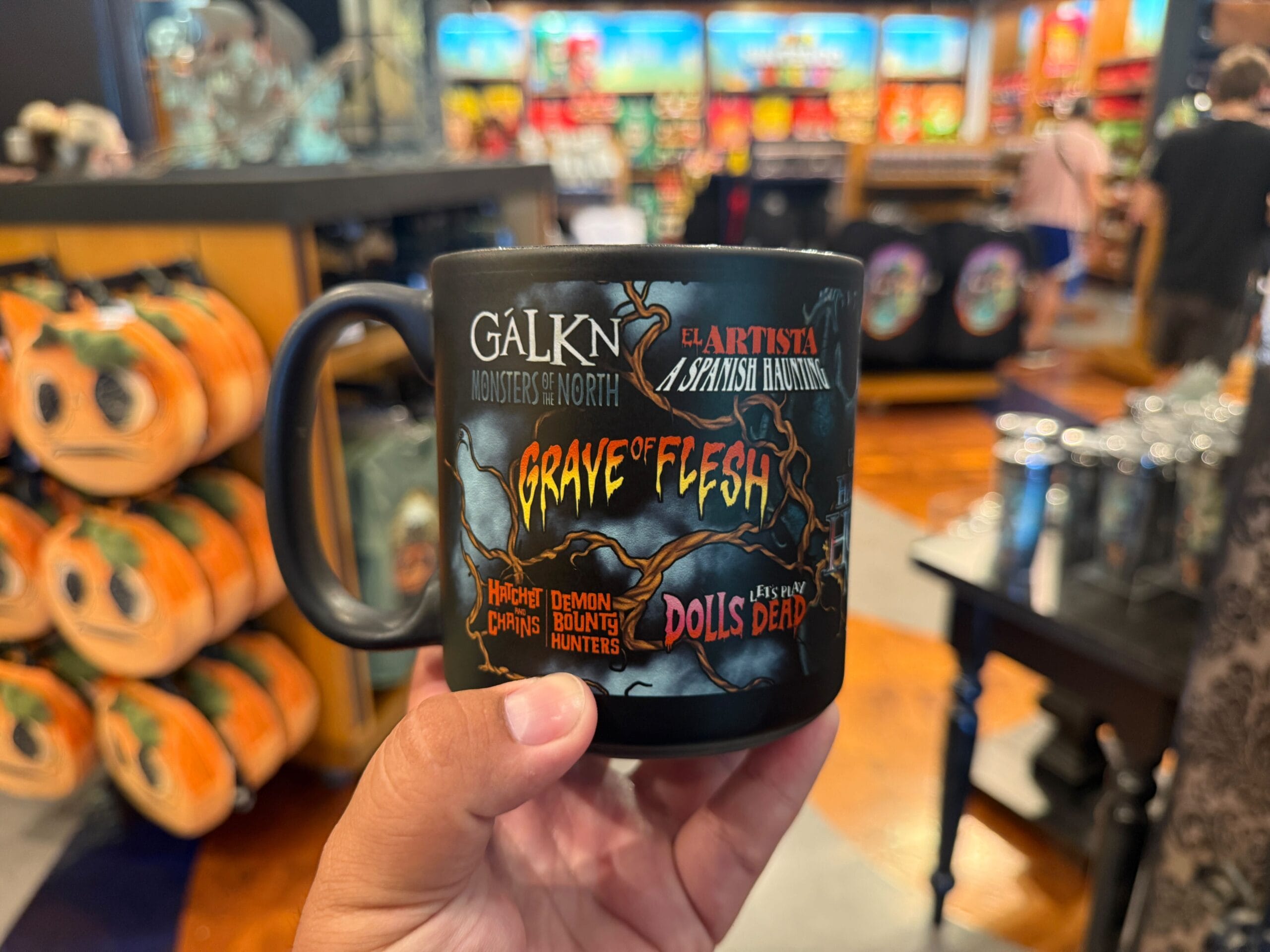A hand holds a black mug with Halloween Horror Nights Merchandise inside a colorful gift shop.