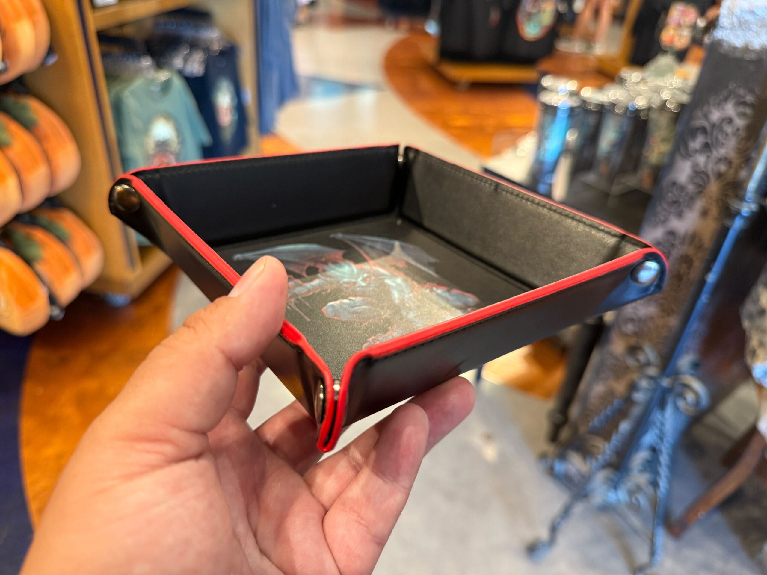 A hand holds a black and red tray with a fantasy dragon, displayed among Halloween Horror Nights Merchandise in-store.