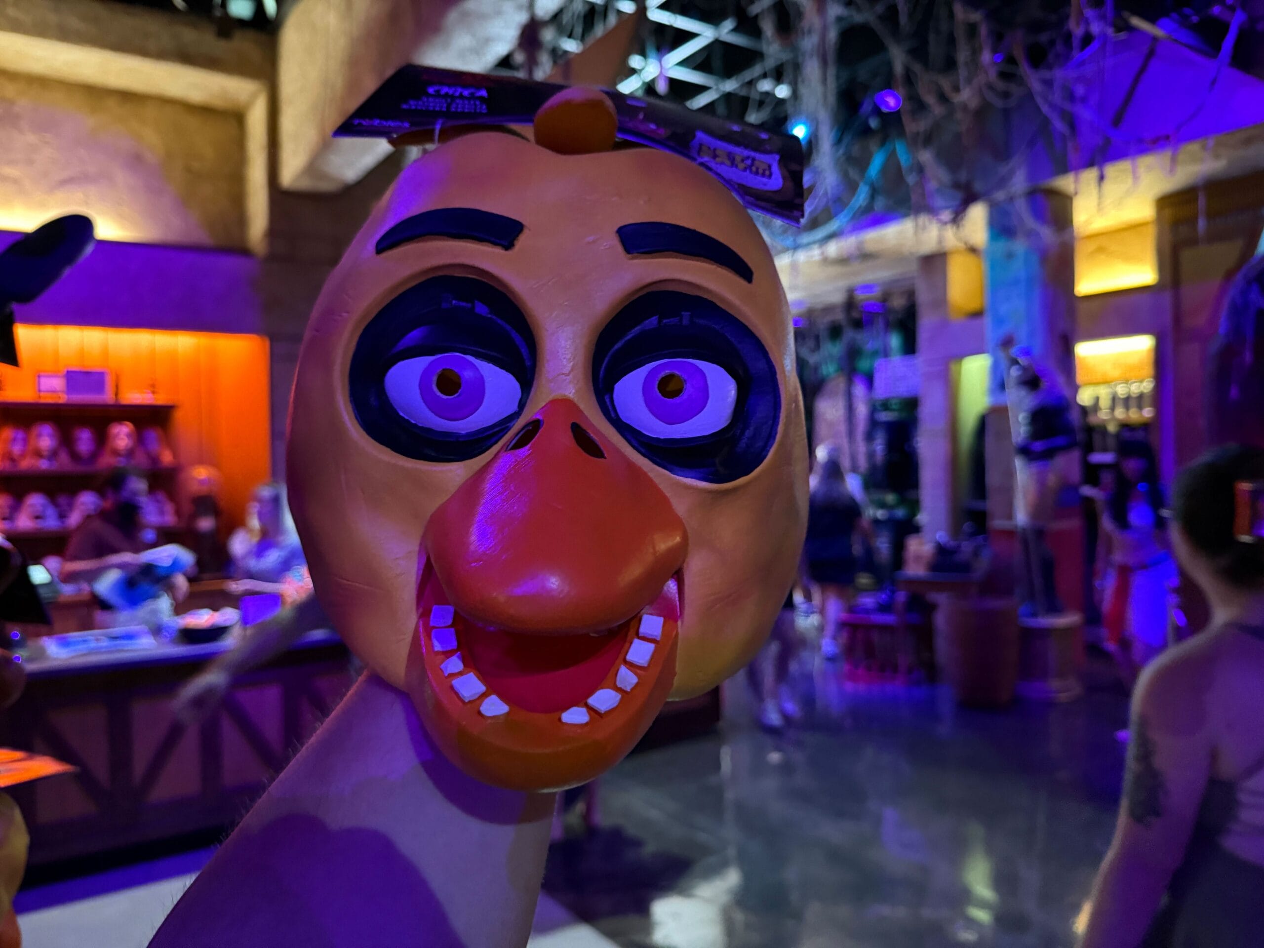 A person holds up a cartoonish orange chicken mask in a dimly lit, colorful store or event space.