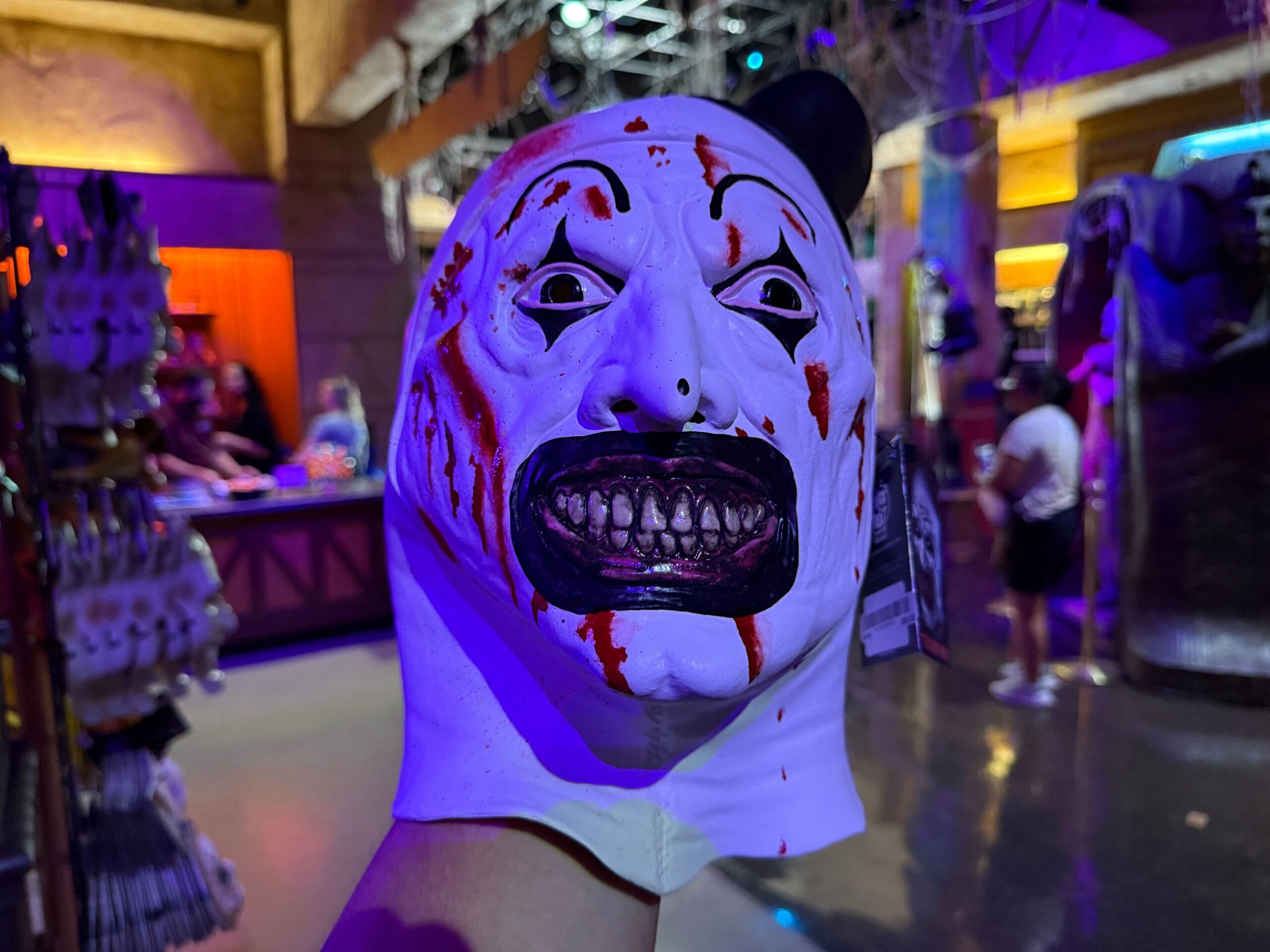 A person holds a creepy, white, blood-splattered clown mask with dark, exaggerated teeth in a brightly lit store.
