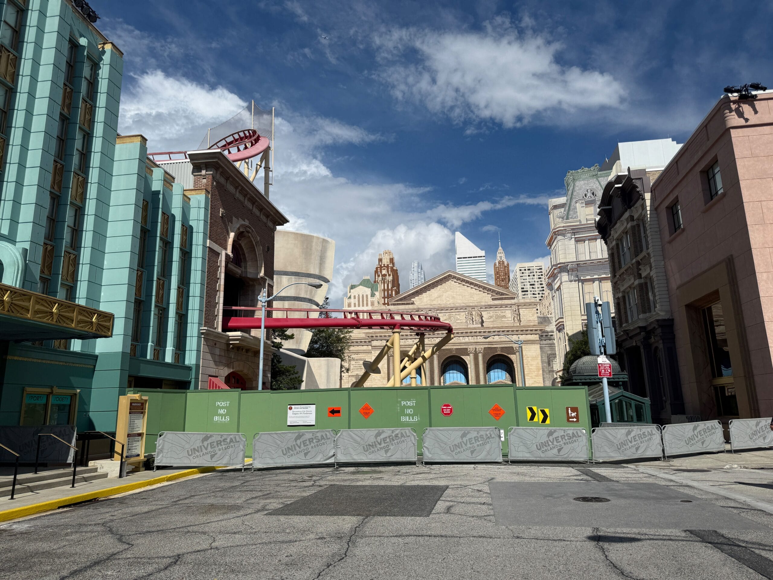 Construction site with green and gray barriers, Rip Ride Rockit track removed, city buildings, and a partly cloudy sky.