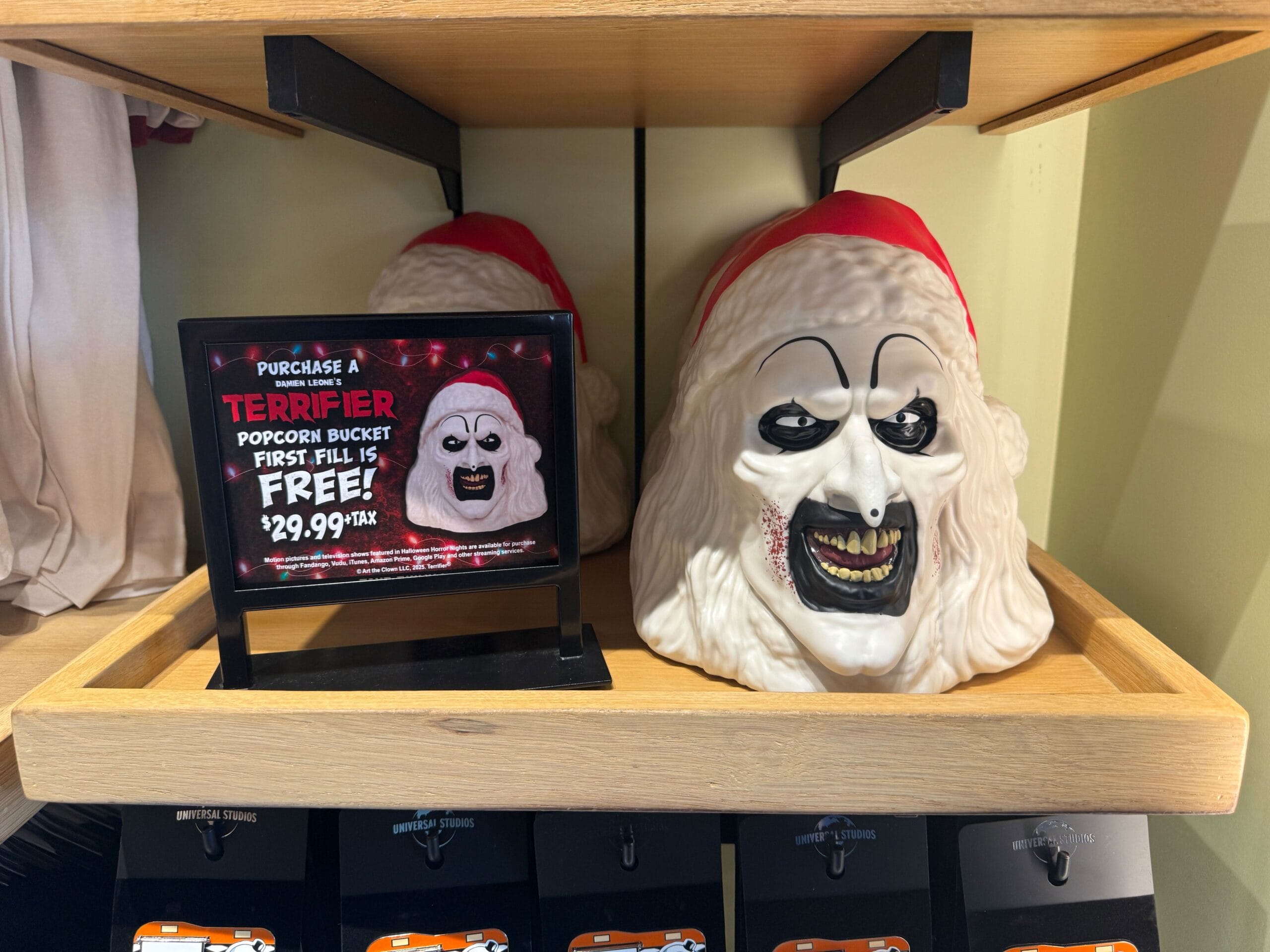 A scary clown face popcorn bucket with a Santa hat is displayed on a shelf next to a sign advertising its sale.