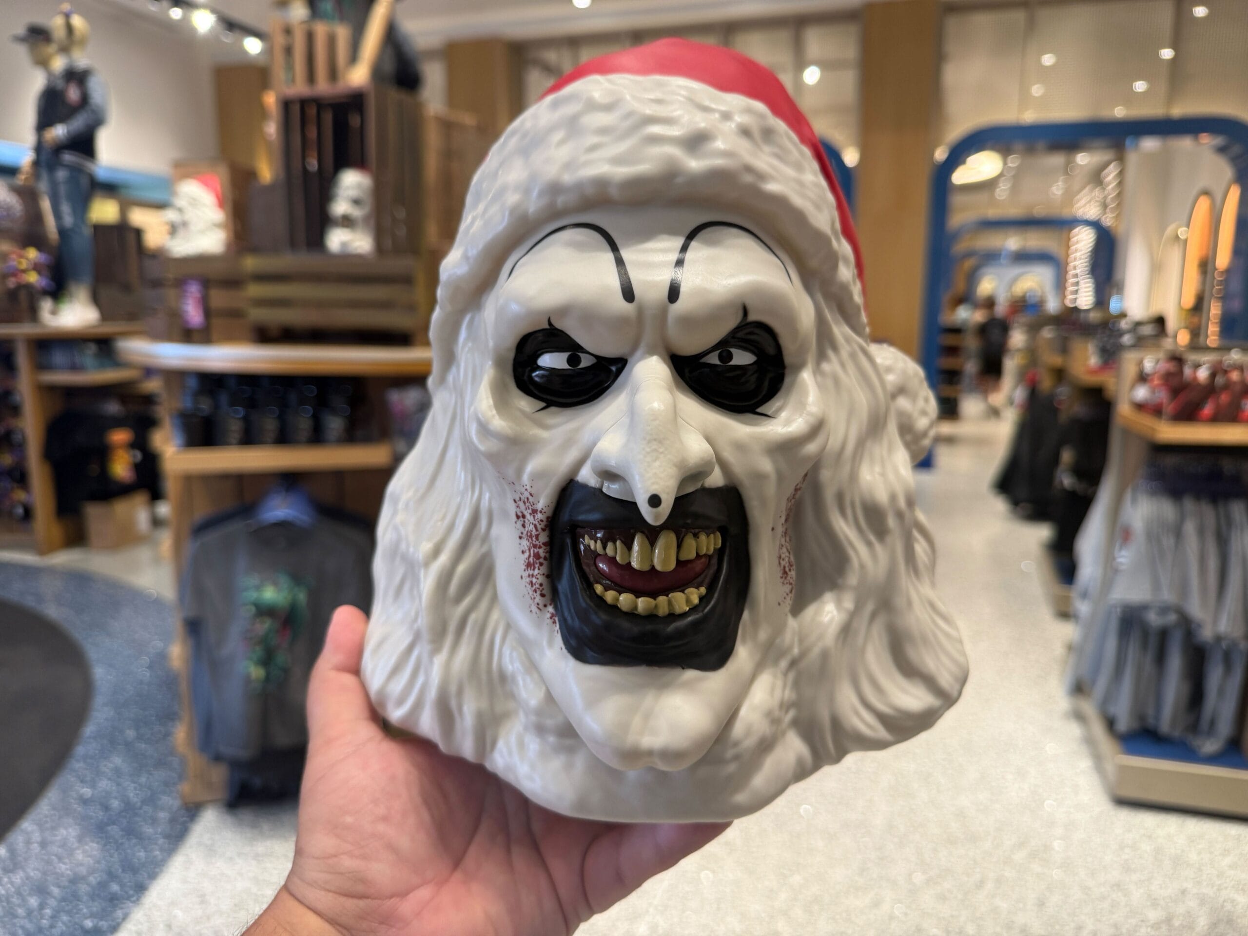 A hand holds a scary white mask with black eyes, sharp teeth, and a Santa hat in a store.