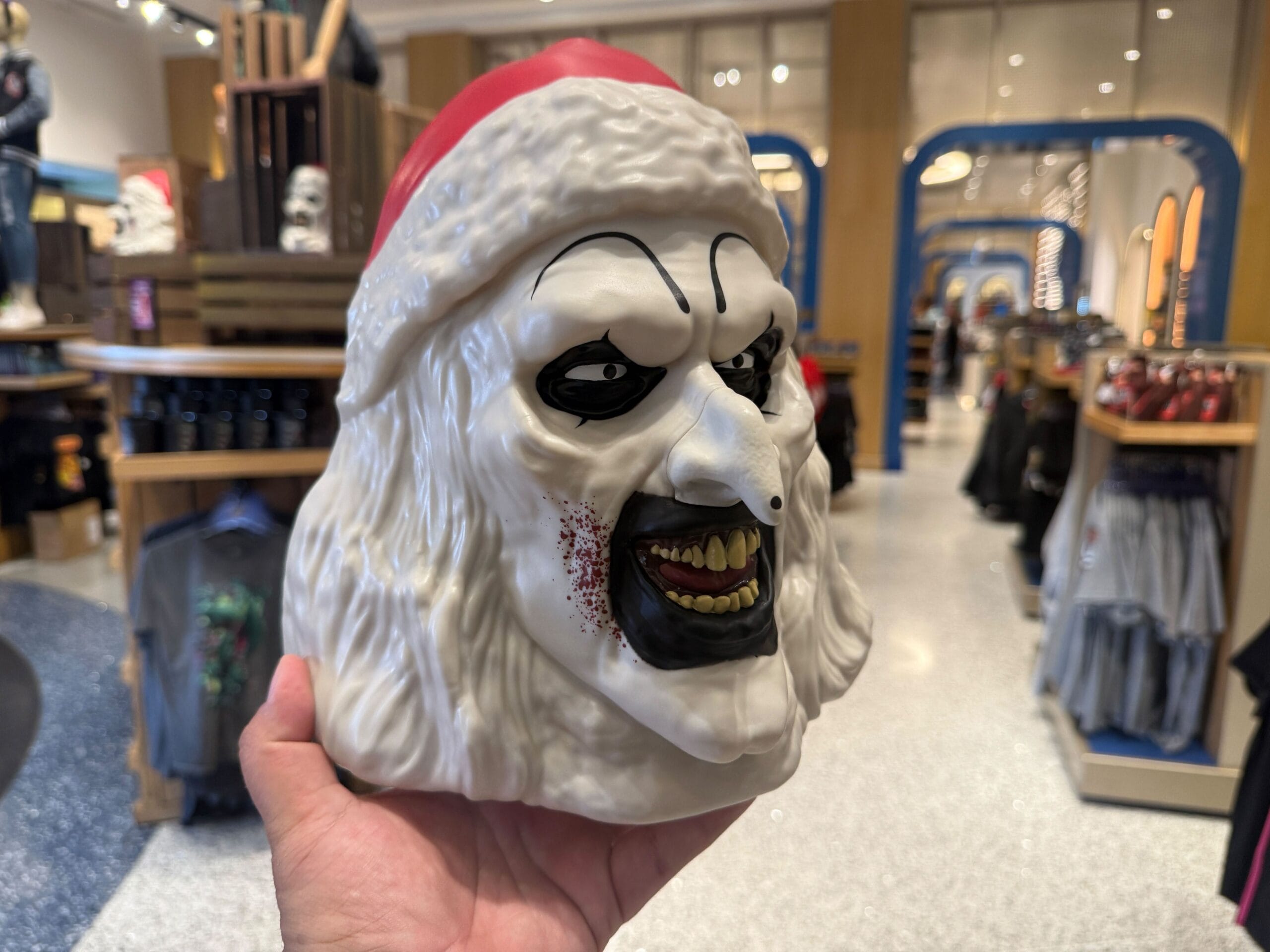 A person holds a creepy Santa Claus mask with white hair, black eye makeup, and bloody teeth in a store.