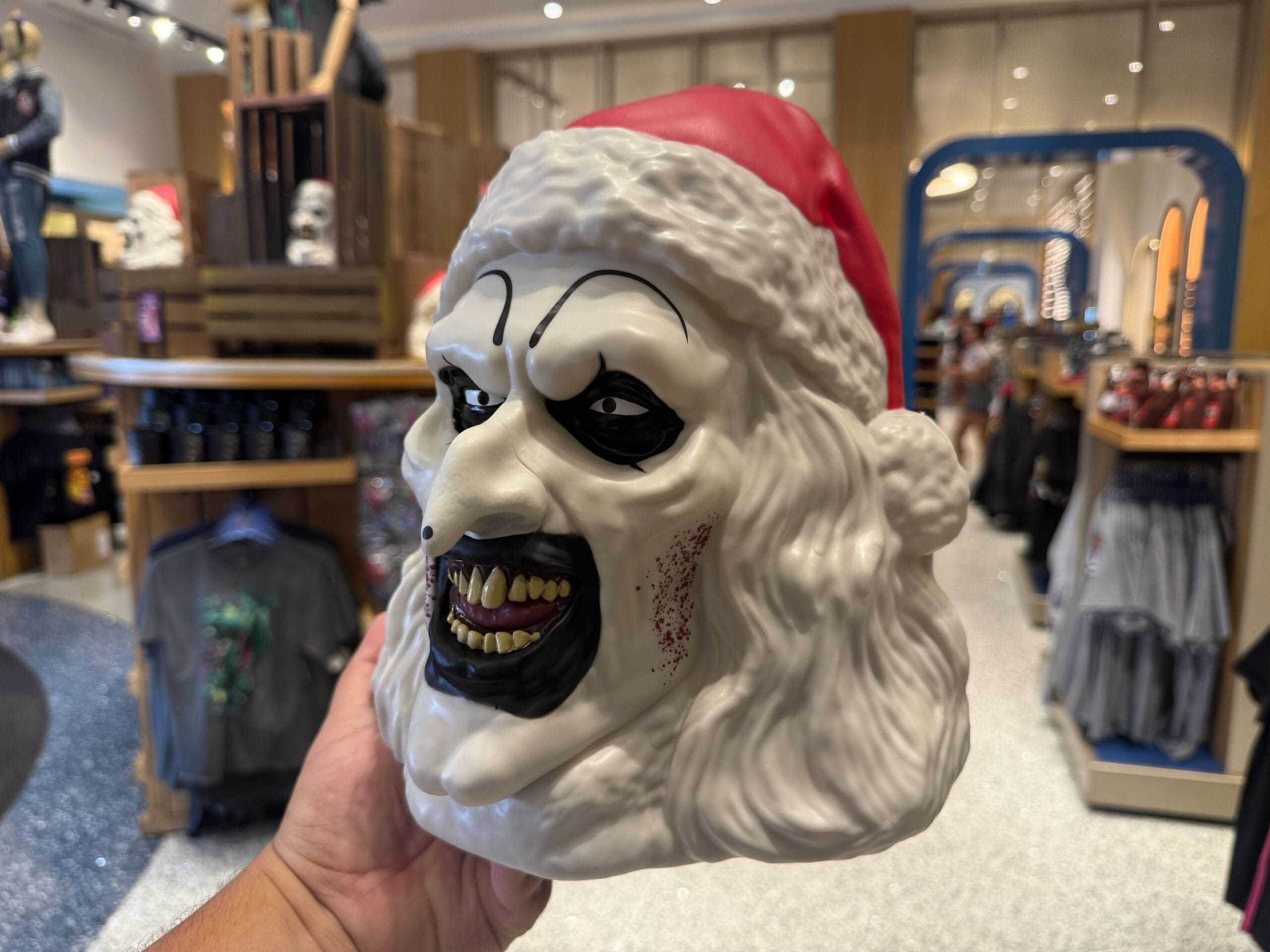 A person holds a scary Santa Claus mask with white hair, sharp teeth, and black eye makeup in a store.