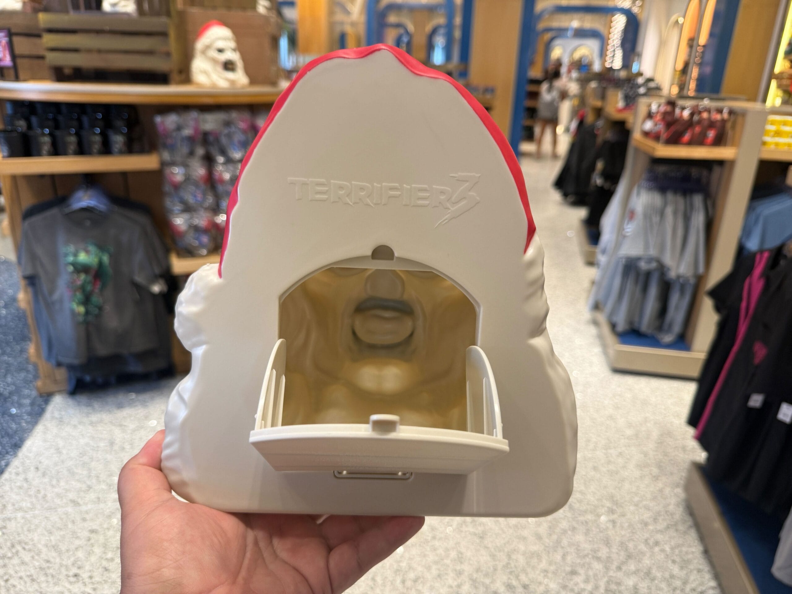 A hand holds a "Terrifier 3" themed popcorn bucket with an open mouth compartment in a store.