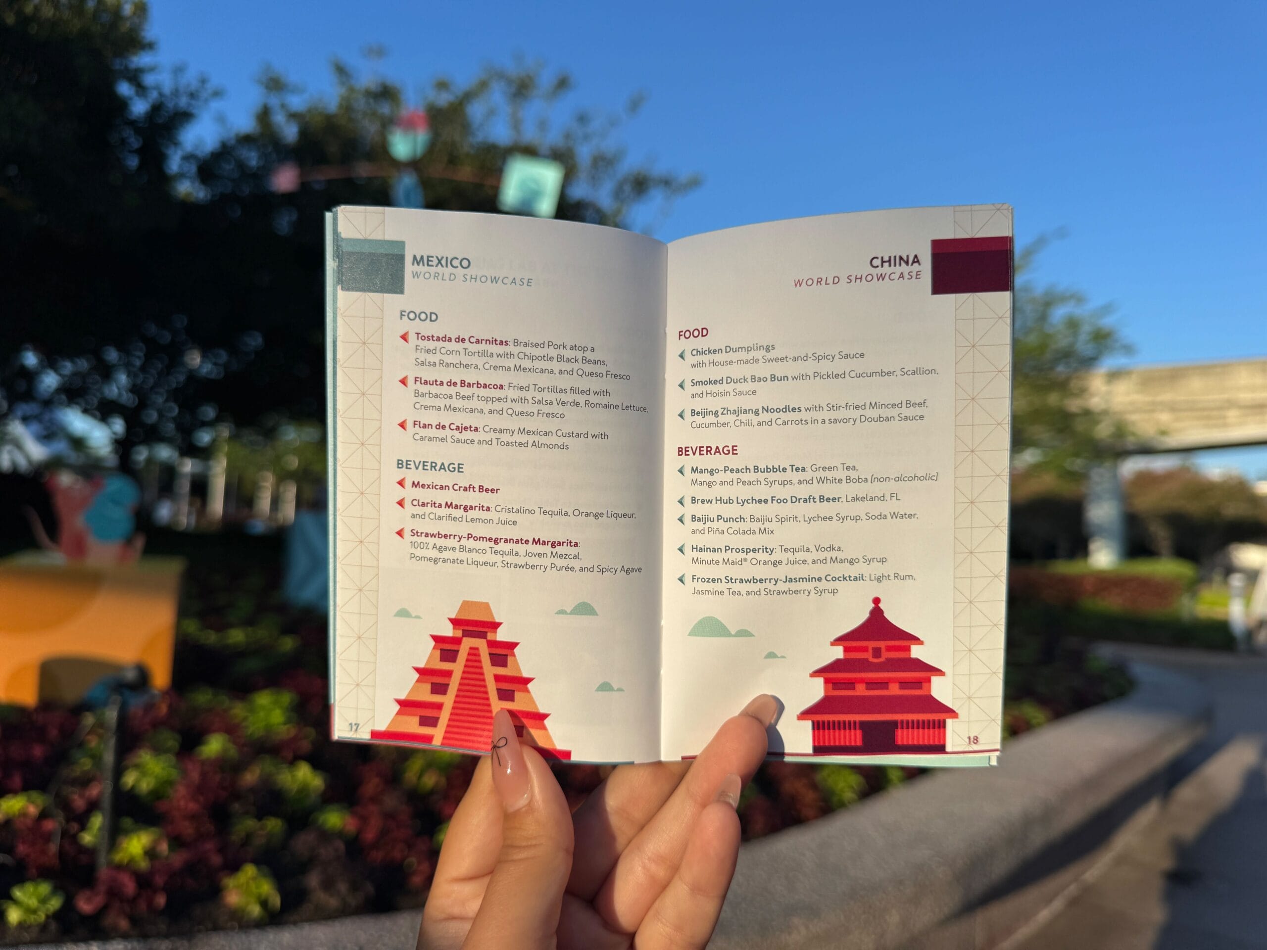 A hand holds an open festival food menu with Mexico and China pavilions listed, outdoors with blue sky and greenery.