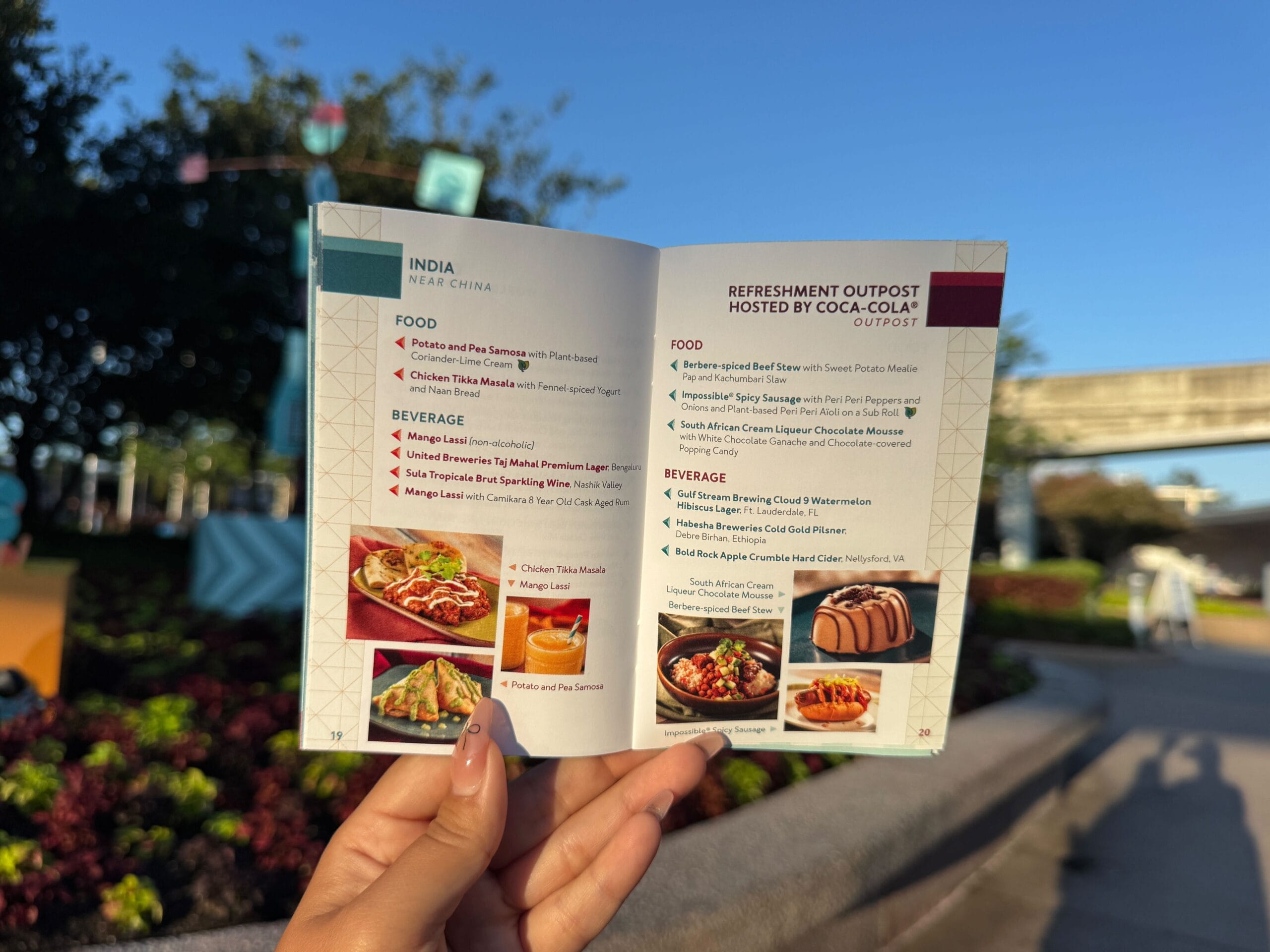 A hand holds an open food festival menu booklet outdoors, with trees and a monorail visible in the background.