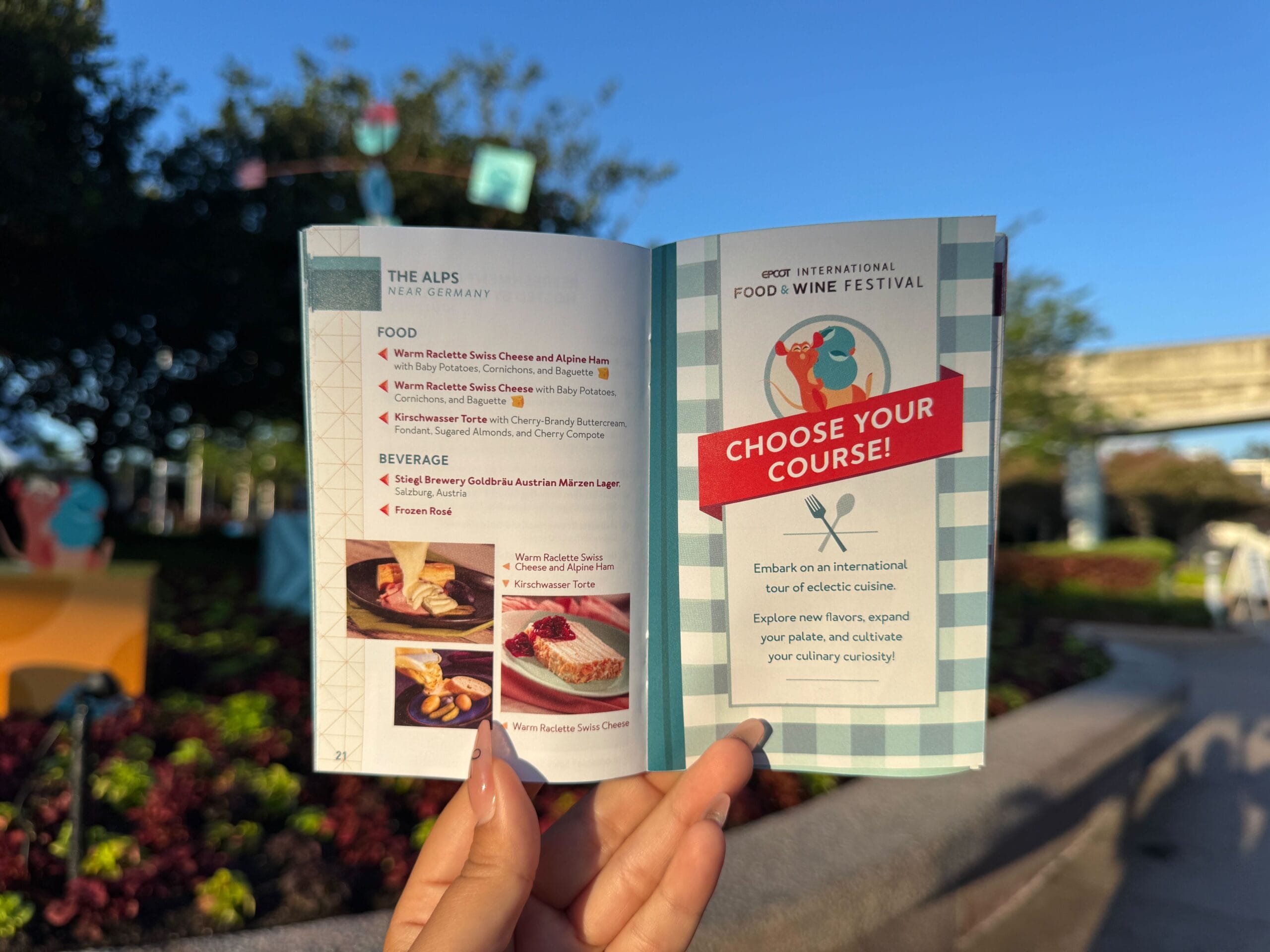 Person holding an open Food & Wine Festival booklet outdoors, with trees and flowers in the background.