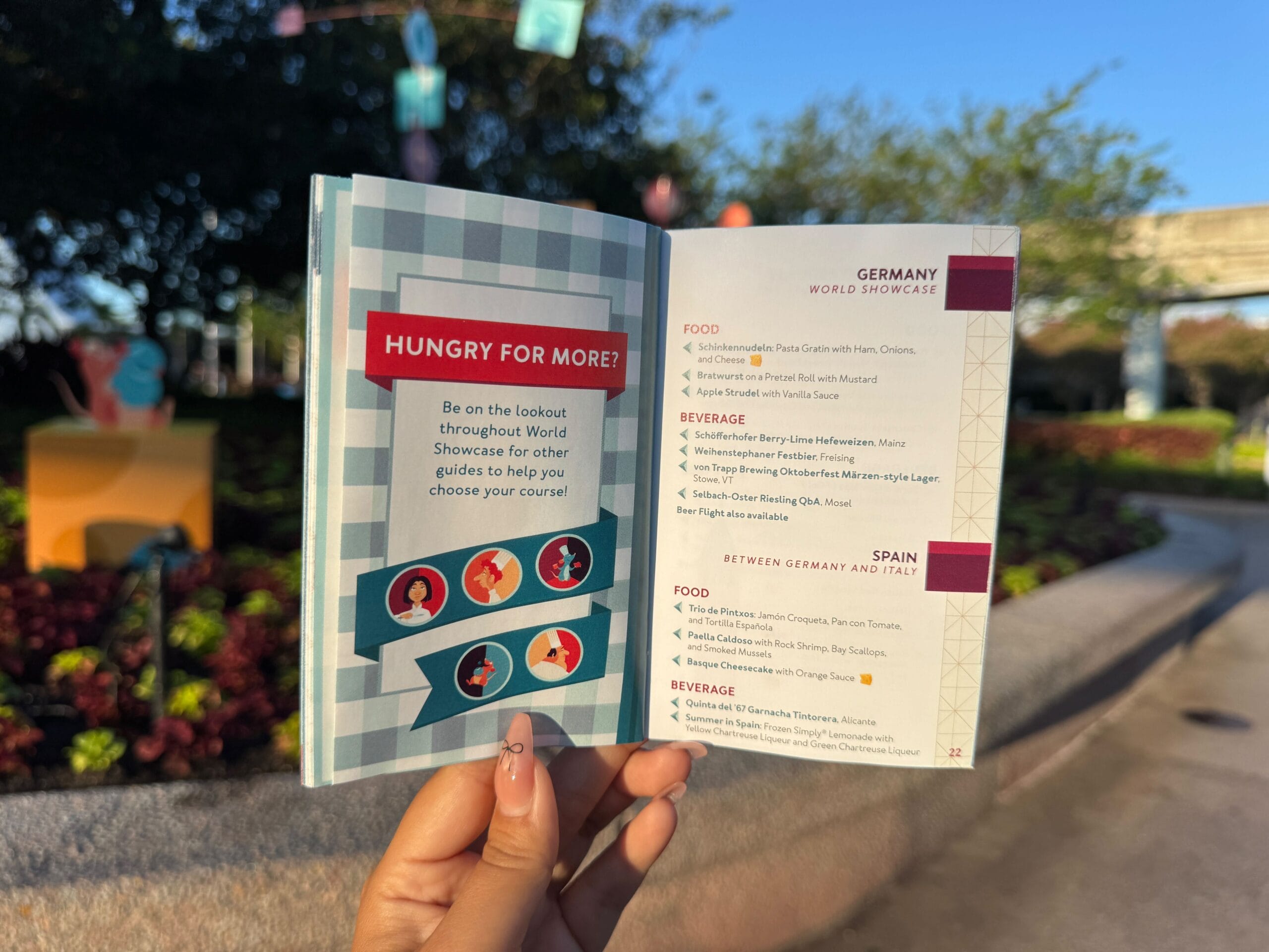A hand holds open a food festival guide outdoors, displaying food and drink options for Germany and Spain.