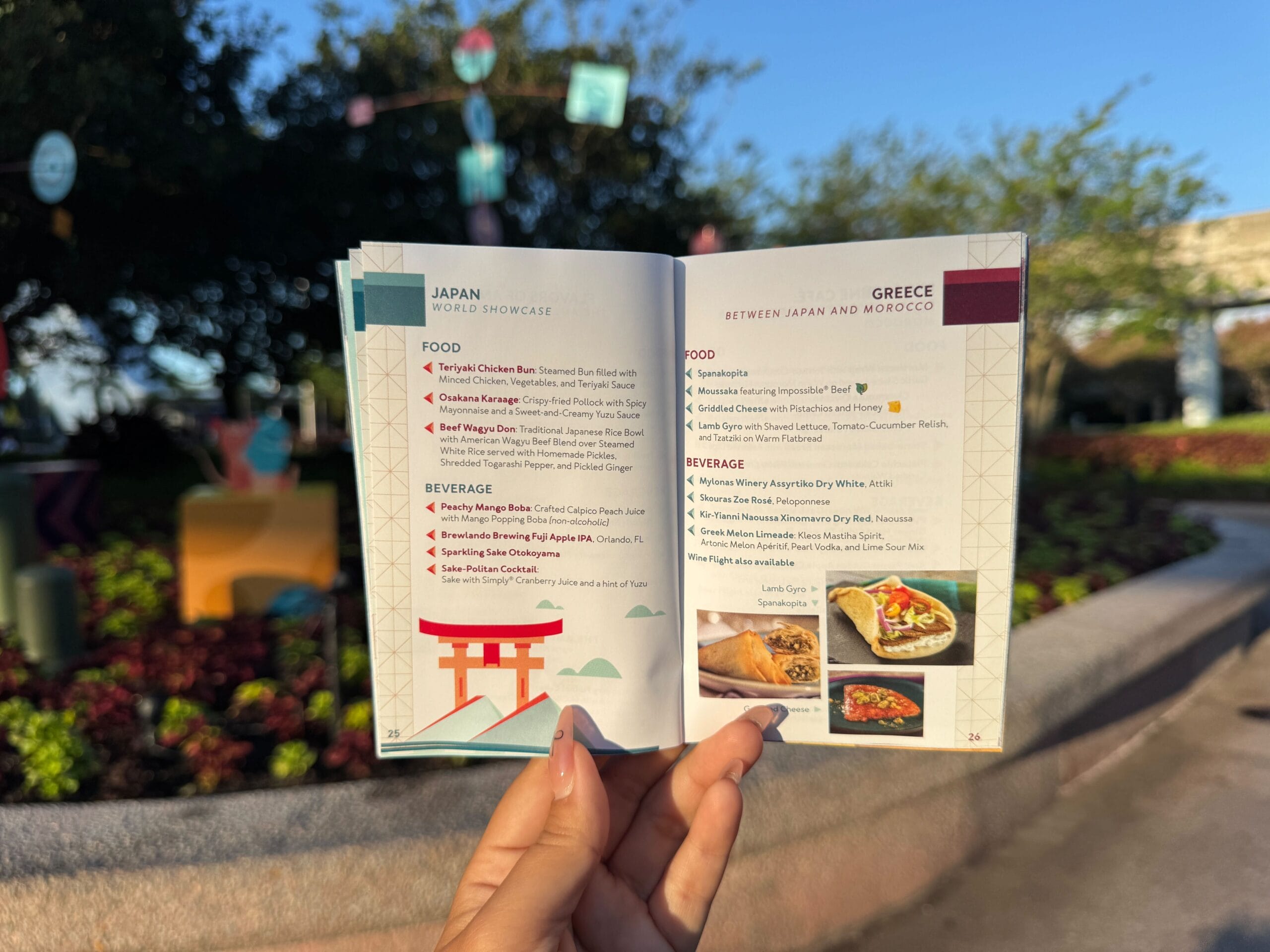 A hand holds a food festival menu booklet open to Japan and Greece pages, with outdoor decor in the background.