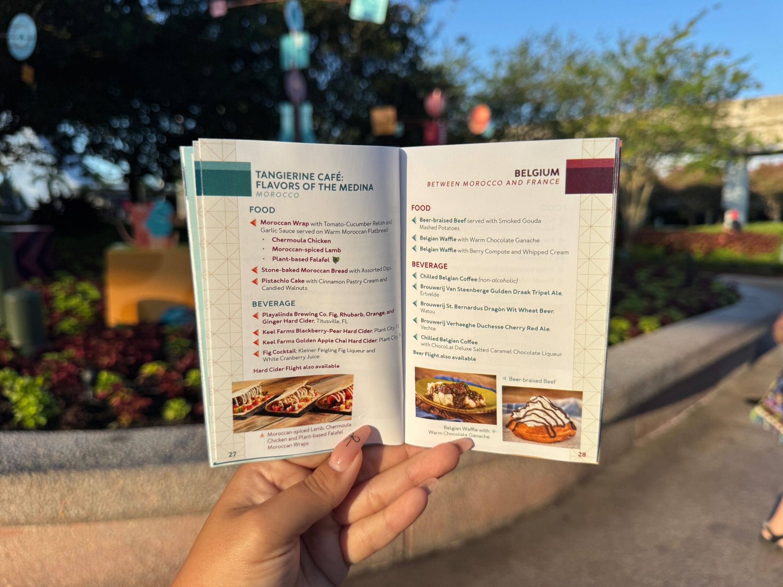A hand holds open a menu booklet outdoors, showing Moroccan and Belgian food and drink options with pictures.