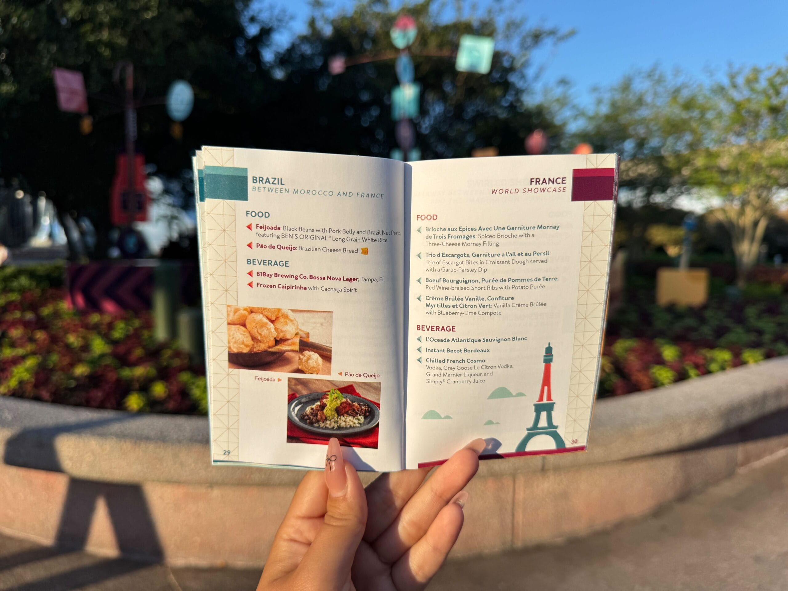 Person holding an open festival food guide outdoors, showing Brazil and France menus with photos of pastries.