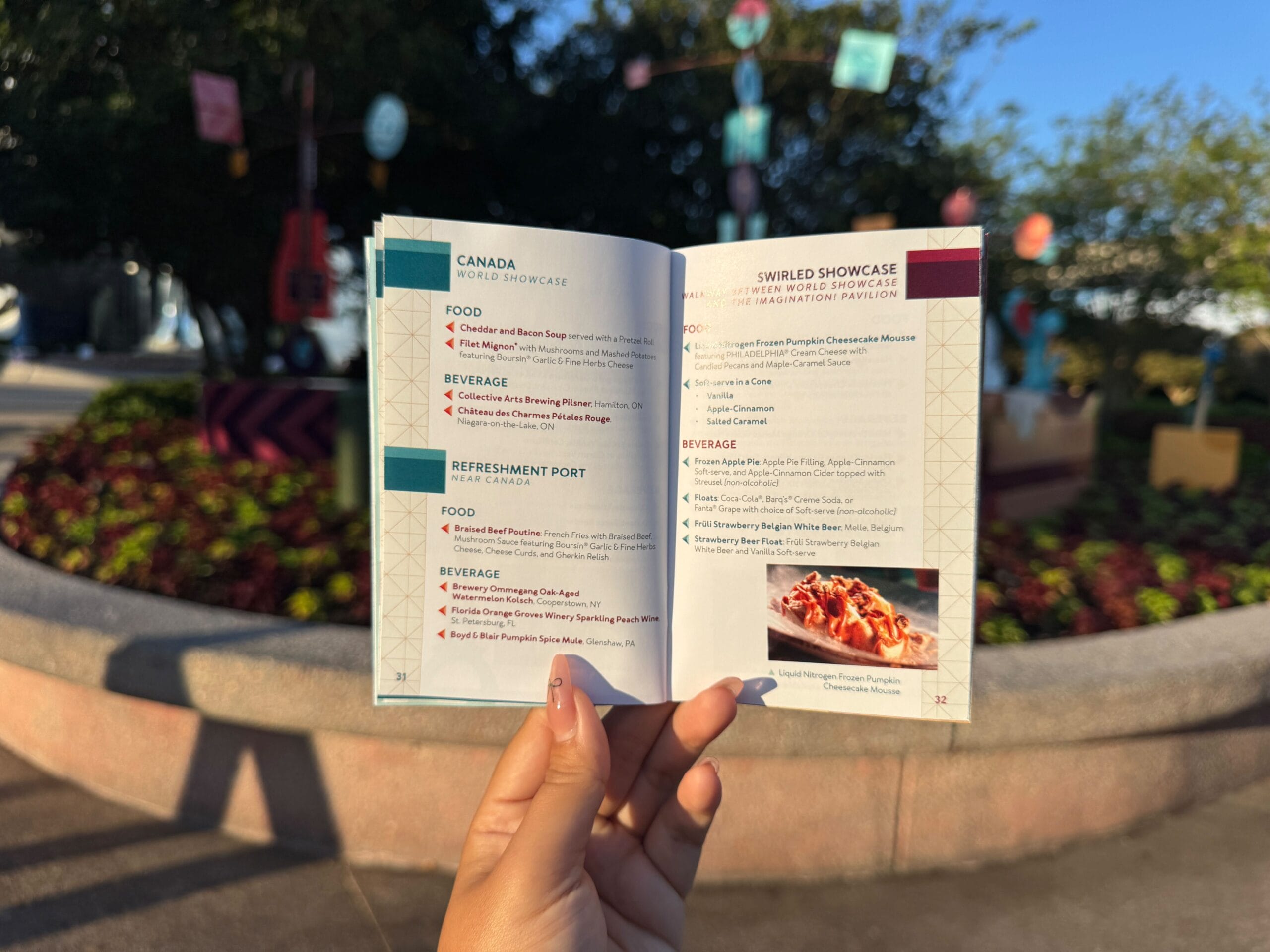 A hand holds open a festival food and beverage menu booklet outdoors in bright sunlight near a planter.