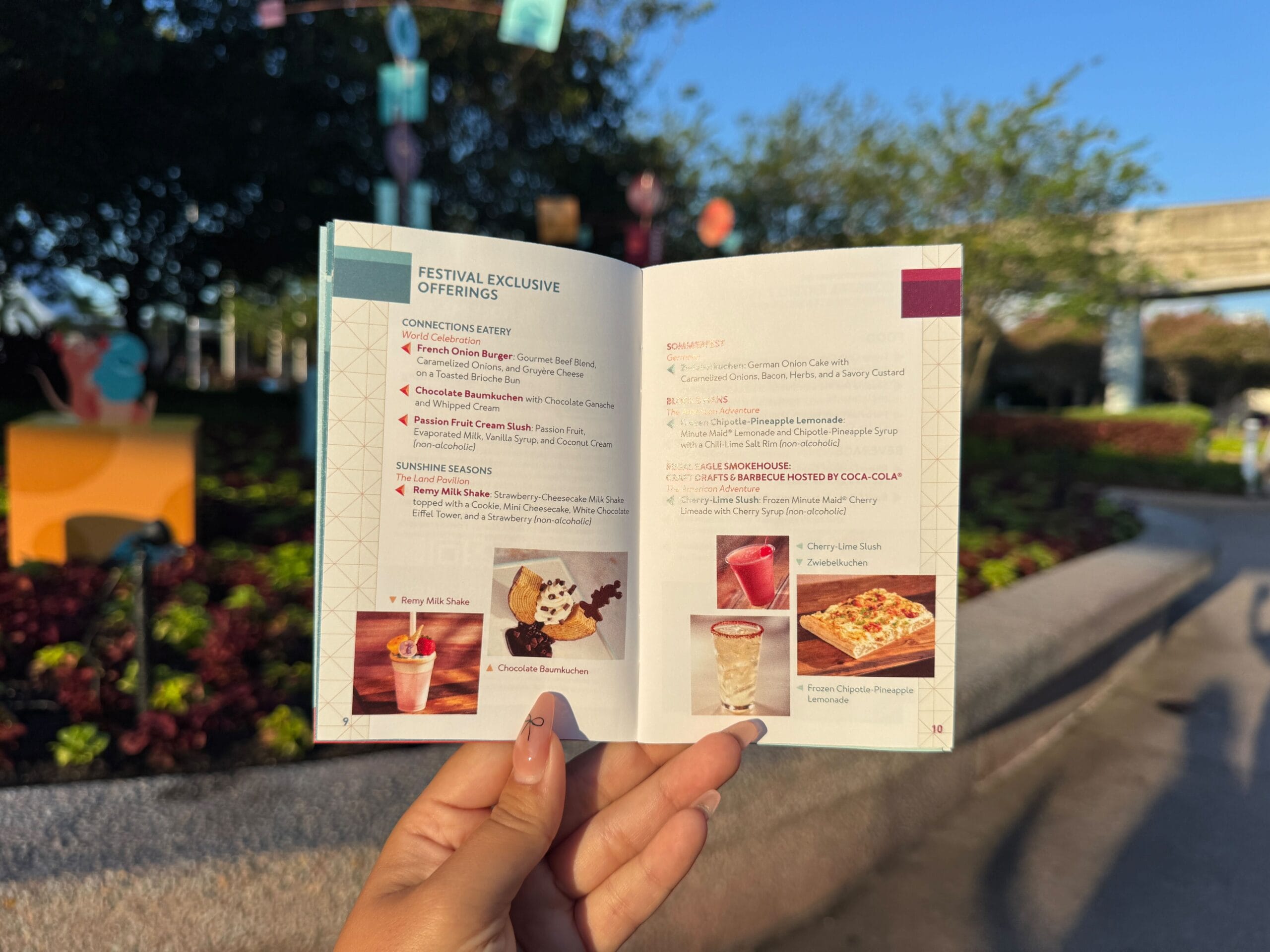 Person holds open a festival menu booklet outdoors, showing food and drink options with colorful decorations in background.