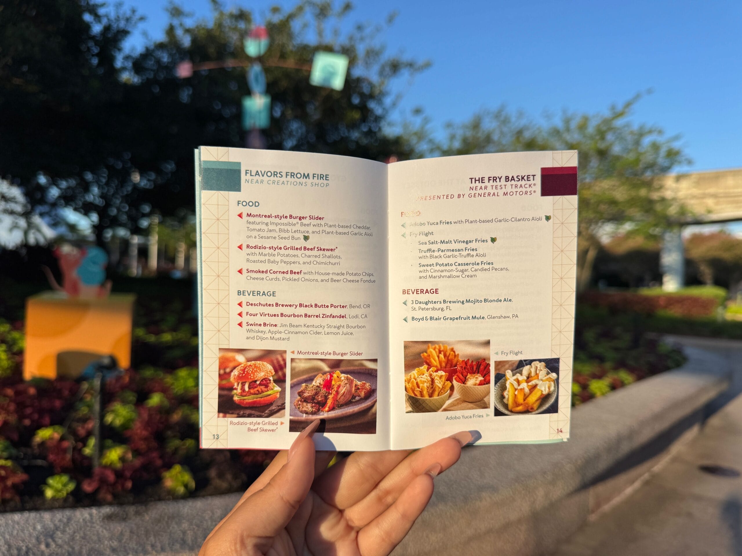 A hand holds an open food and beverage menu outdoors with trees and decorations in the background.