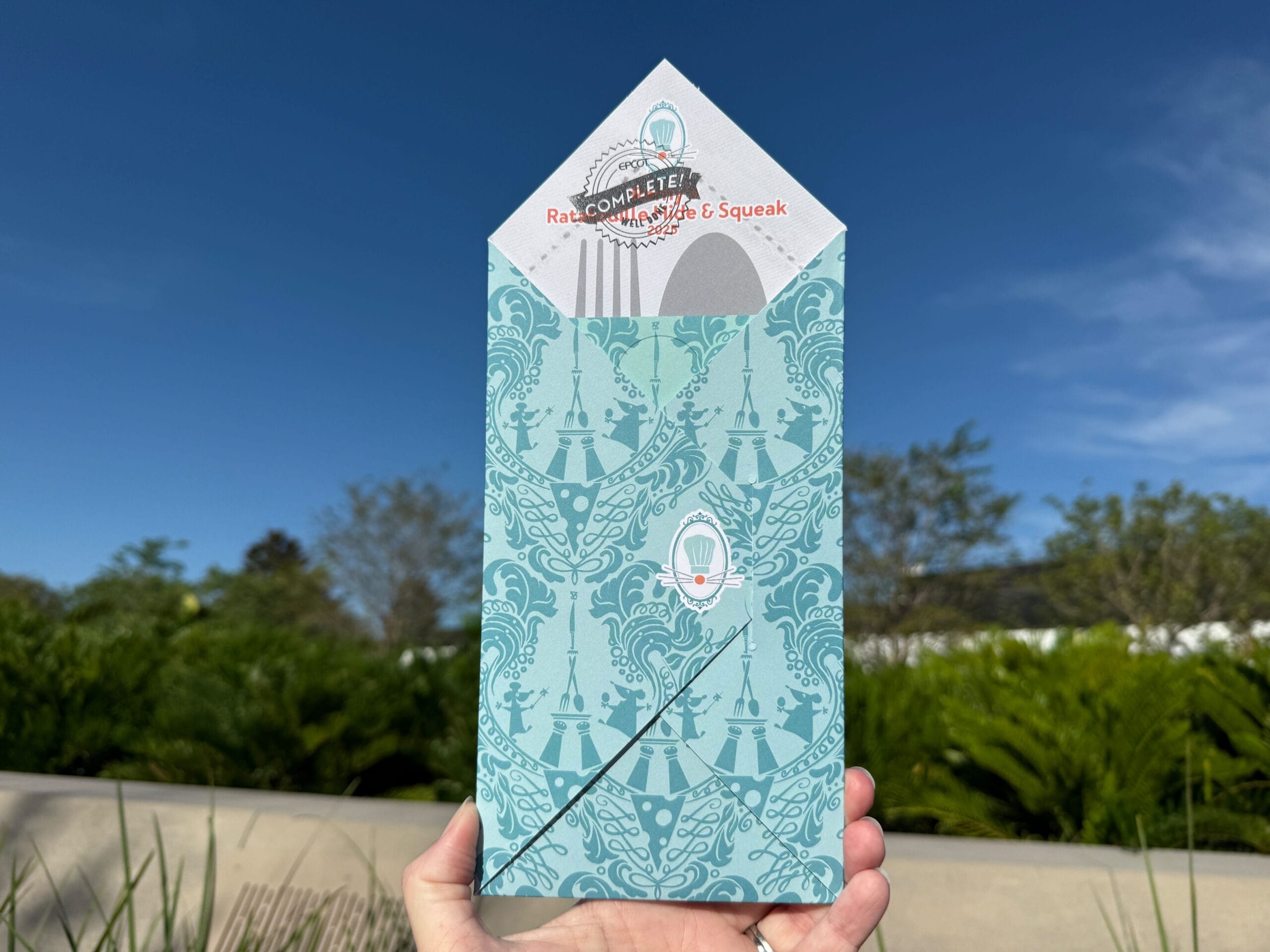 A hand holds a decorative blue envelope from EPCOT Food & Wine Festival’s Remy's Ratatouille Hide & Squeak scavenger hunt.