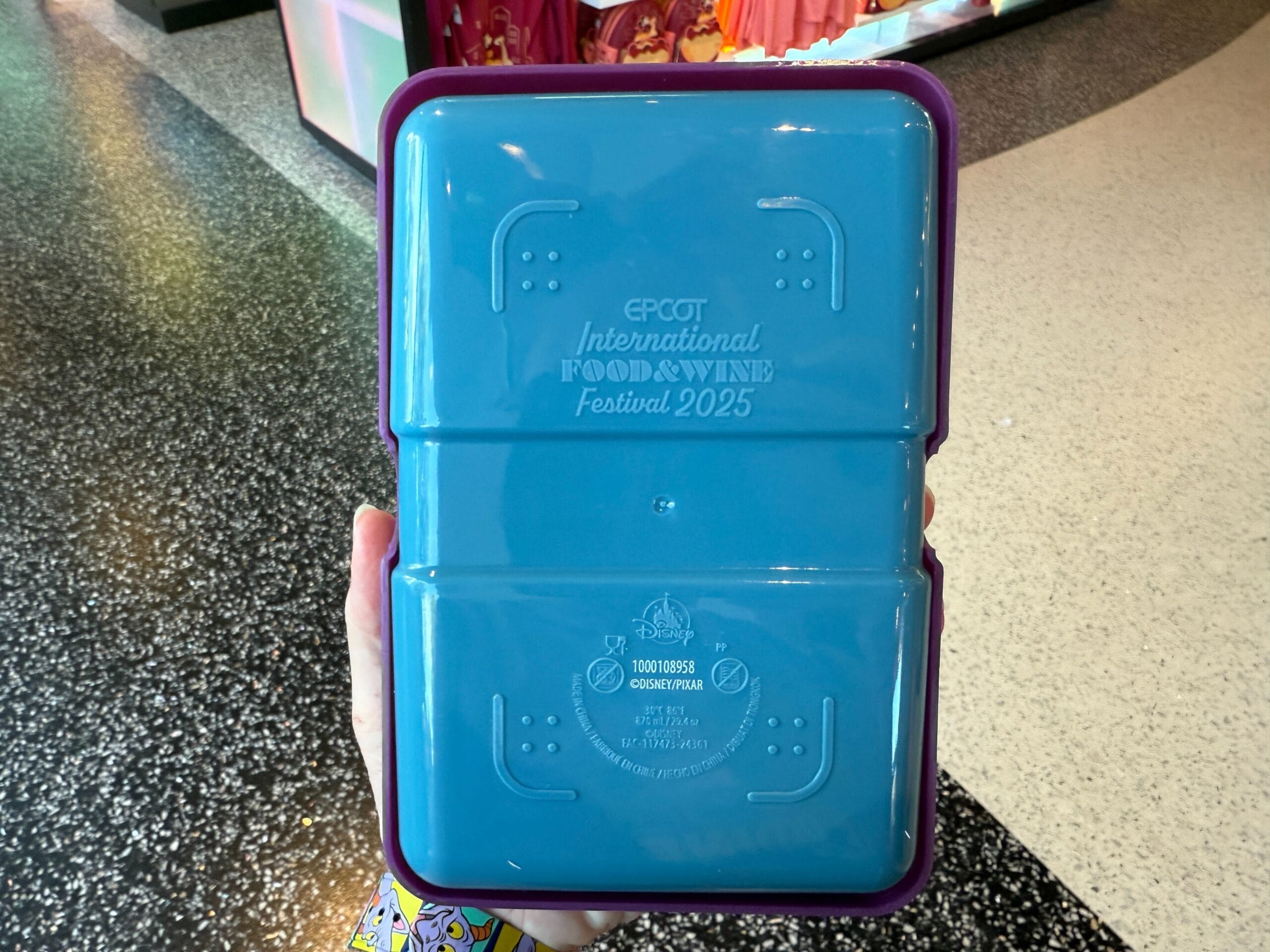 A hand holds a blue EPCOT Food & Wine Festival 2025 container, perfect for Remy's Ratatouille Hide & Squeak scavenger hunt.