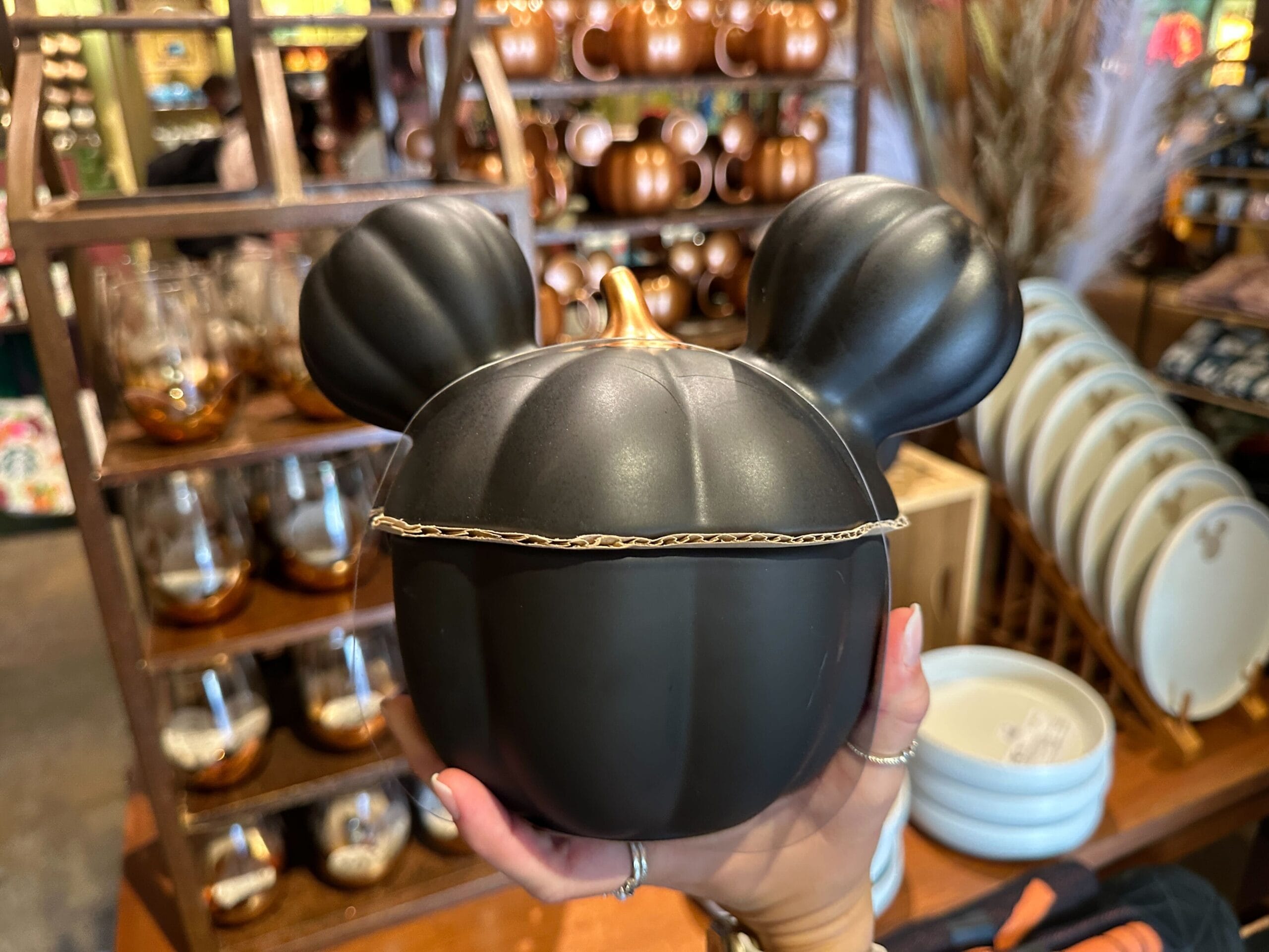 A hand holds a black pumpkin container shaped like a Mickey icon with ears in a store with Animal Kingdom Halloween home decor.