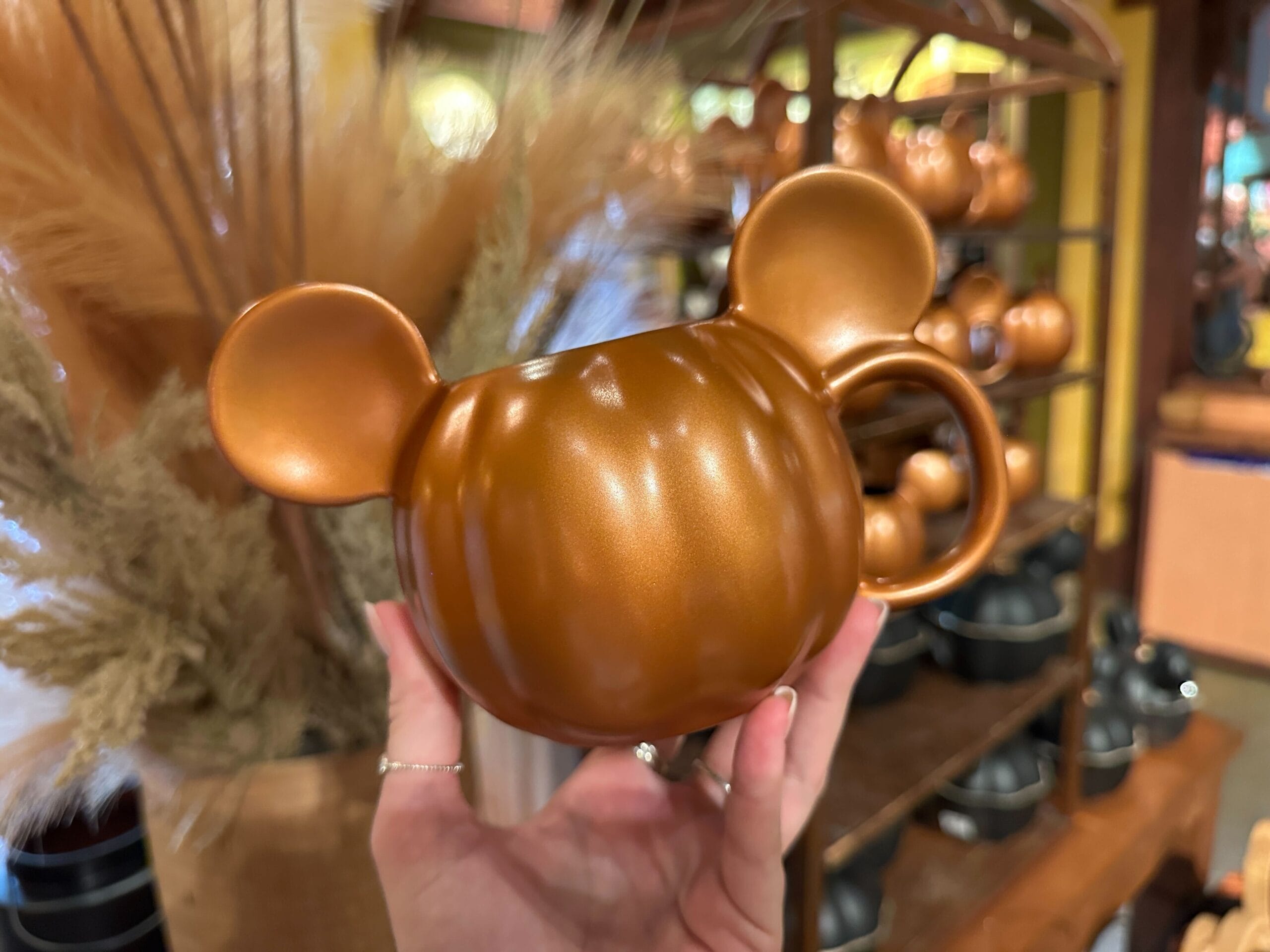 A hand holds a pumpkin-shaped mug with Mickey Mouse ears in a store filled with Halloween home decor.