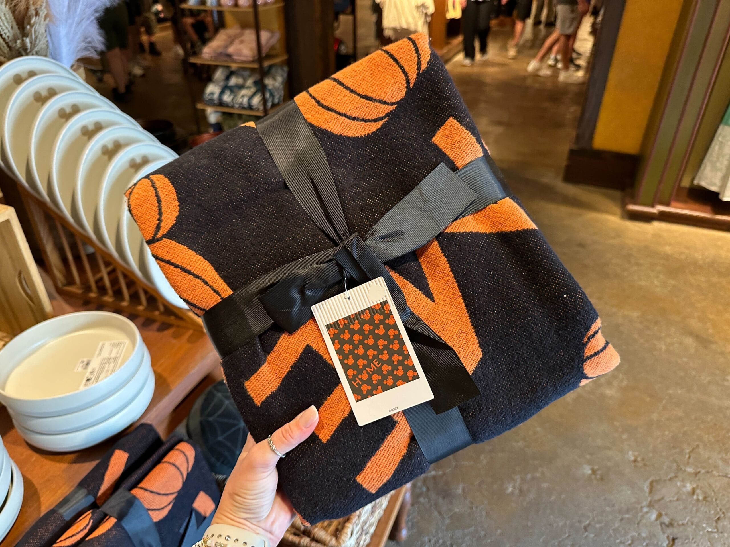 A hand holds a black and orange blanket tied with a ribbon—perfect Halloween Home Decor—in a store with dishes behind.
