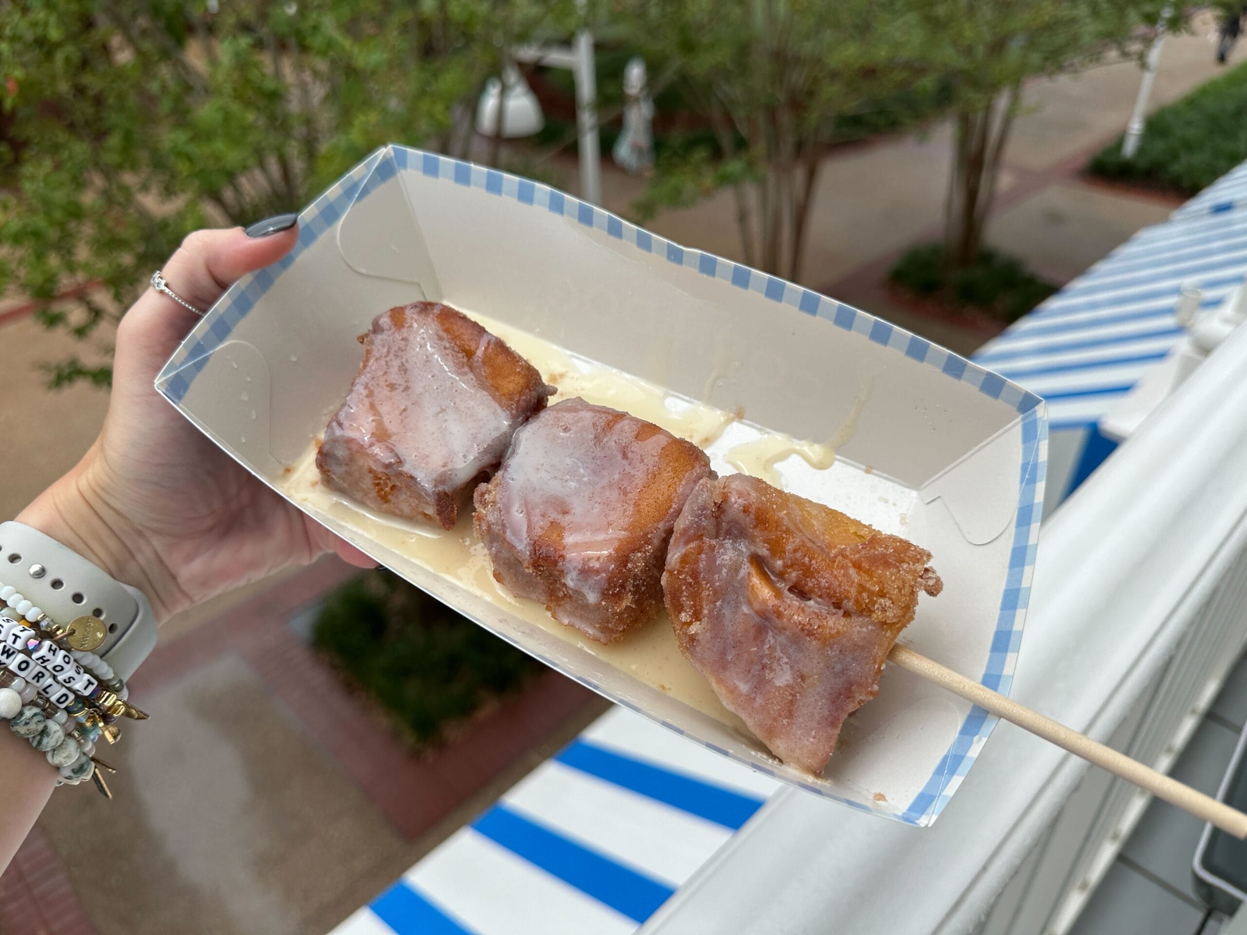 Three pieces of fried Blue Ribbon Corn Dogs on a skewer, topped with icing, held above blue-and-white striped awnings outside.