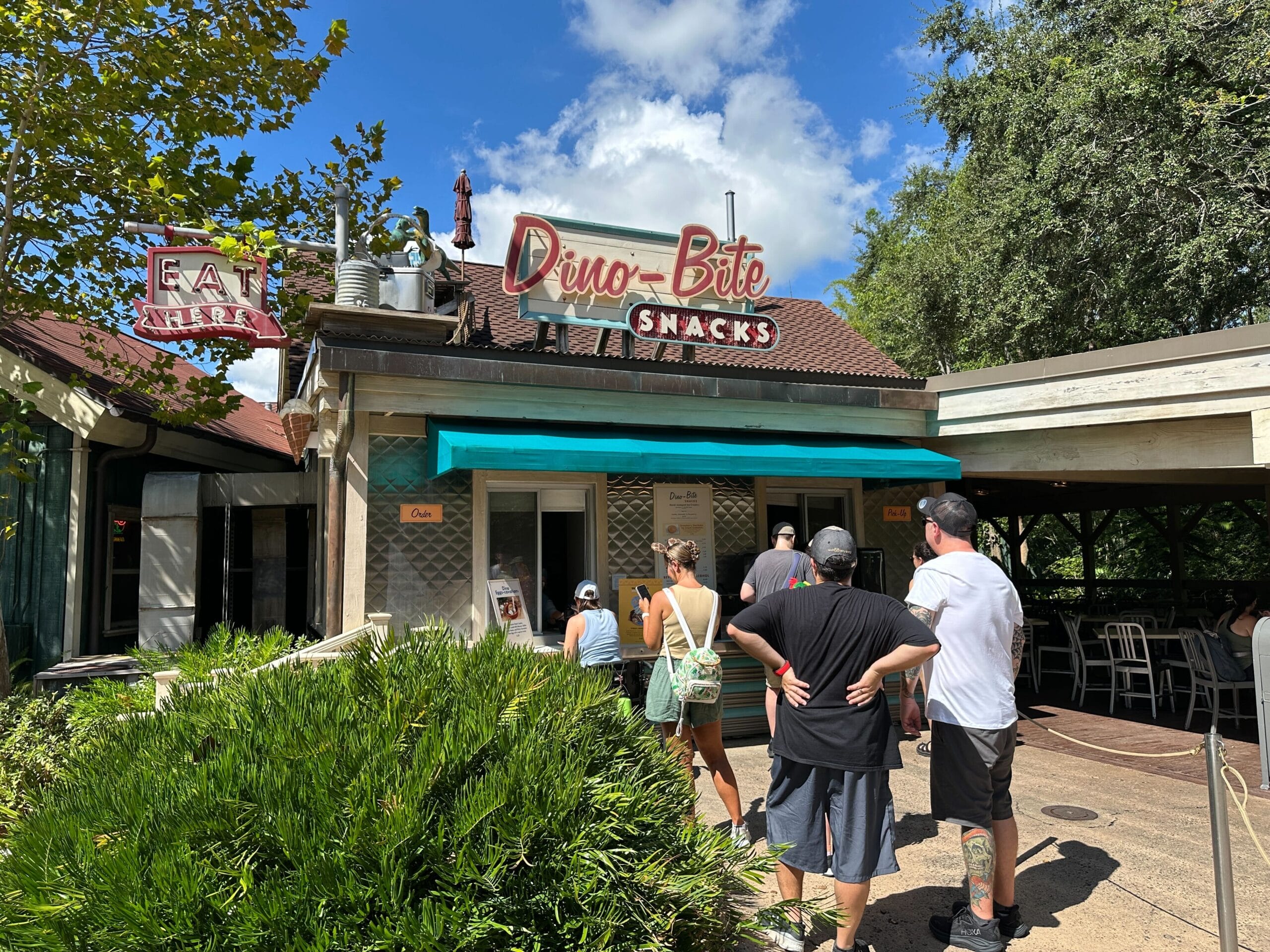 People stand in line outside Dino-Bite Snacks, a snack shop with a vintage sign on a sunny day.