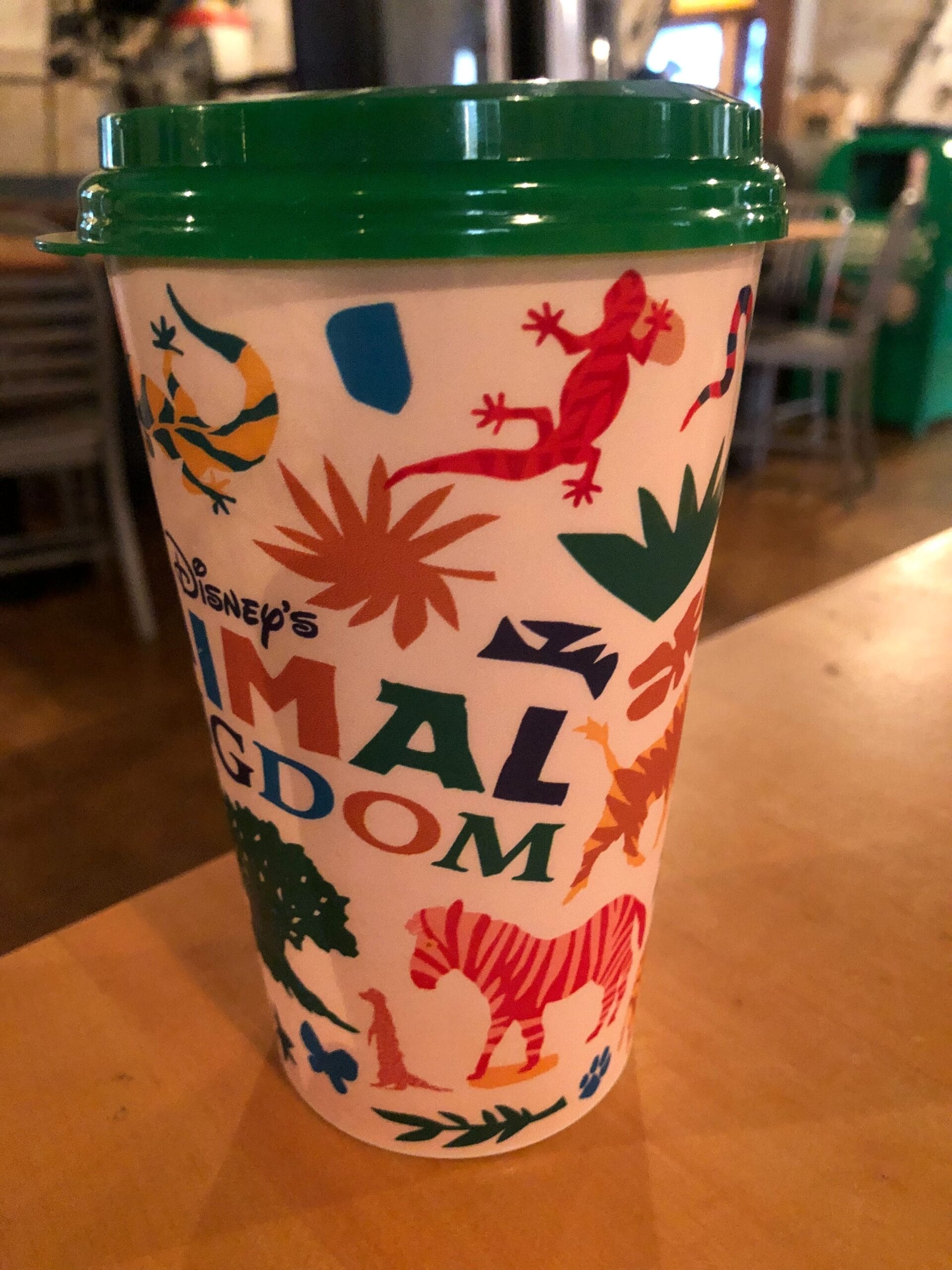 A Disney’s Animal Kingdom cup with colorful animal and plant designs and a green lid sitting on a wooden table.