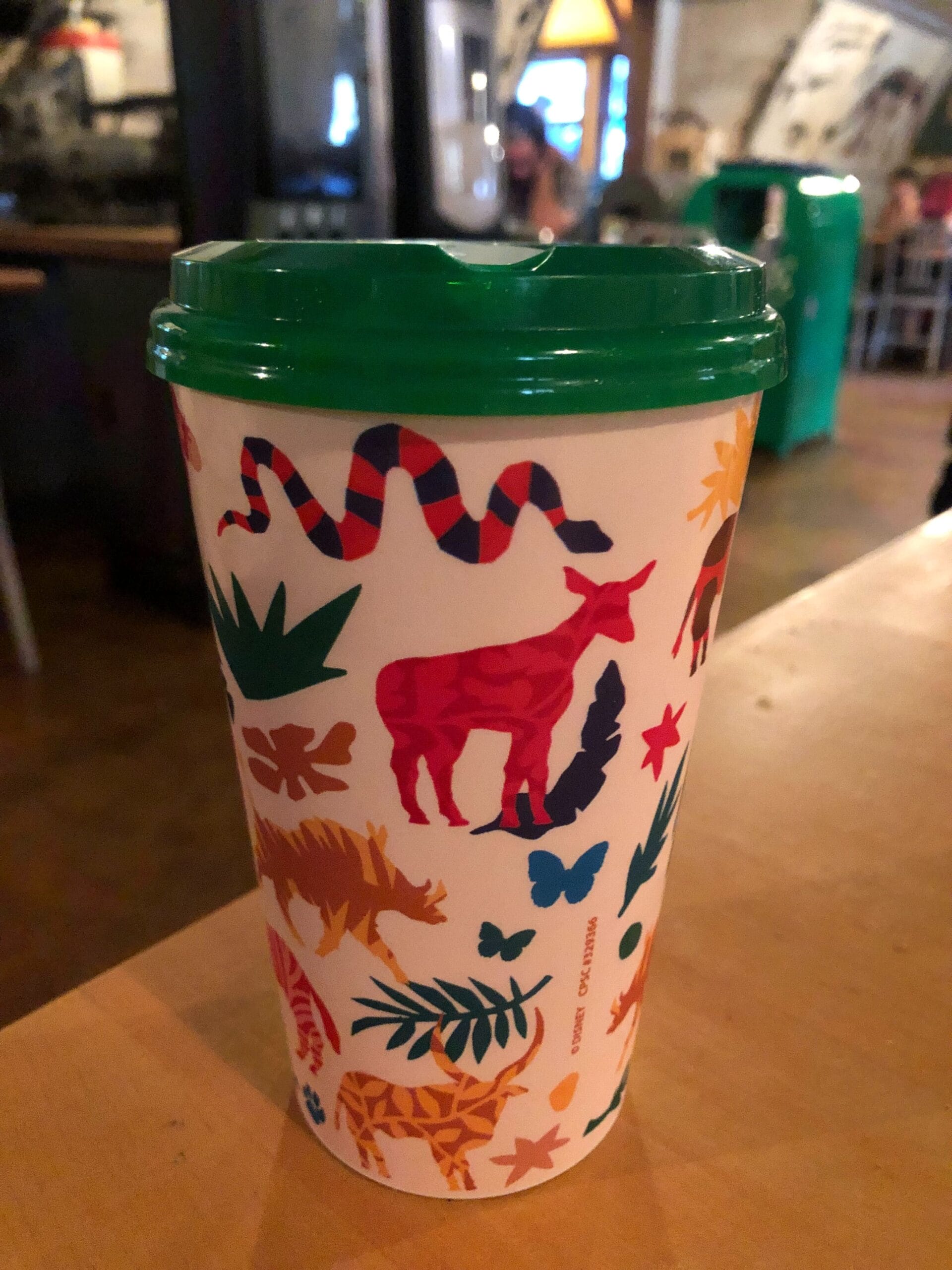 A colorful cup with animal and plant illustrations and a green lid sits on a wooden table indoors.