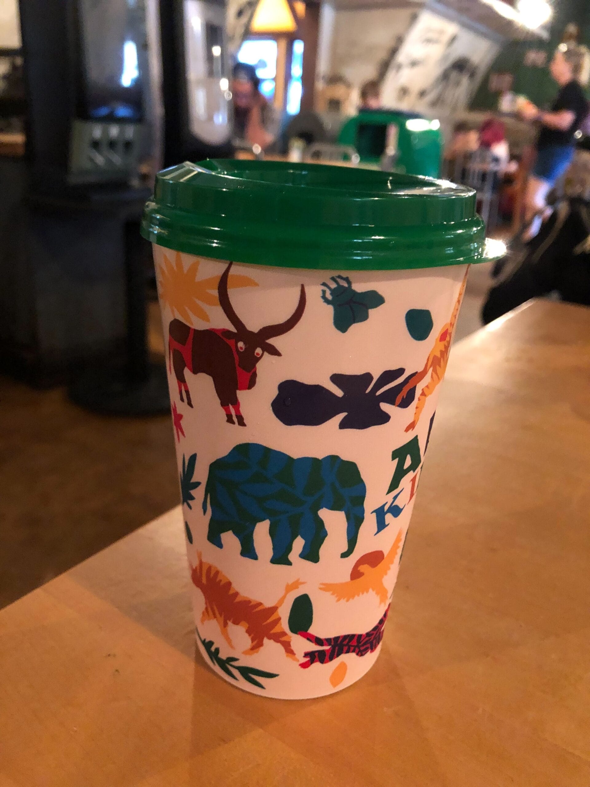 A white cup with a green lid, decorated with colorful animal silhouettes and leaves, sits on a wooden table indoors.