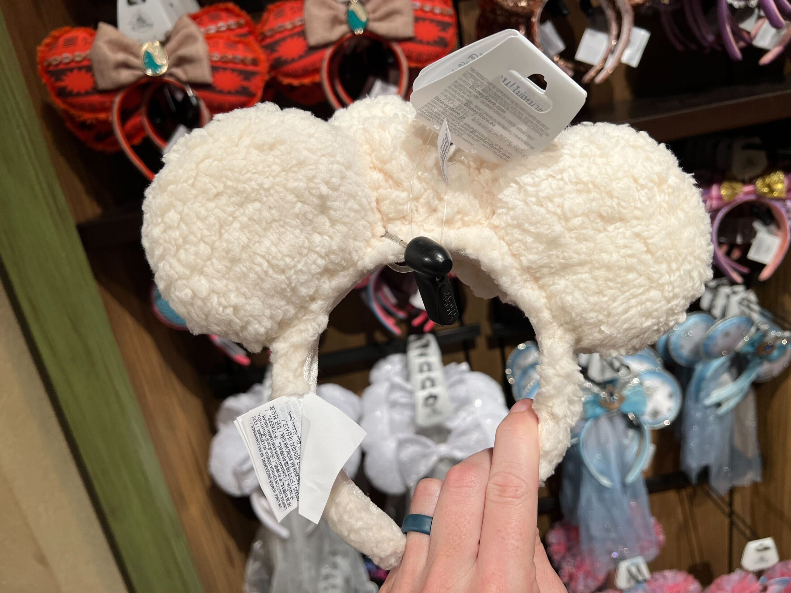 A hand holds a plush ear headband in a store display filled with similar fuzzy, cream-colored options.