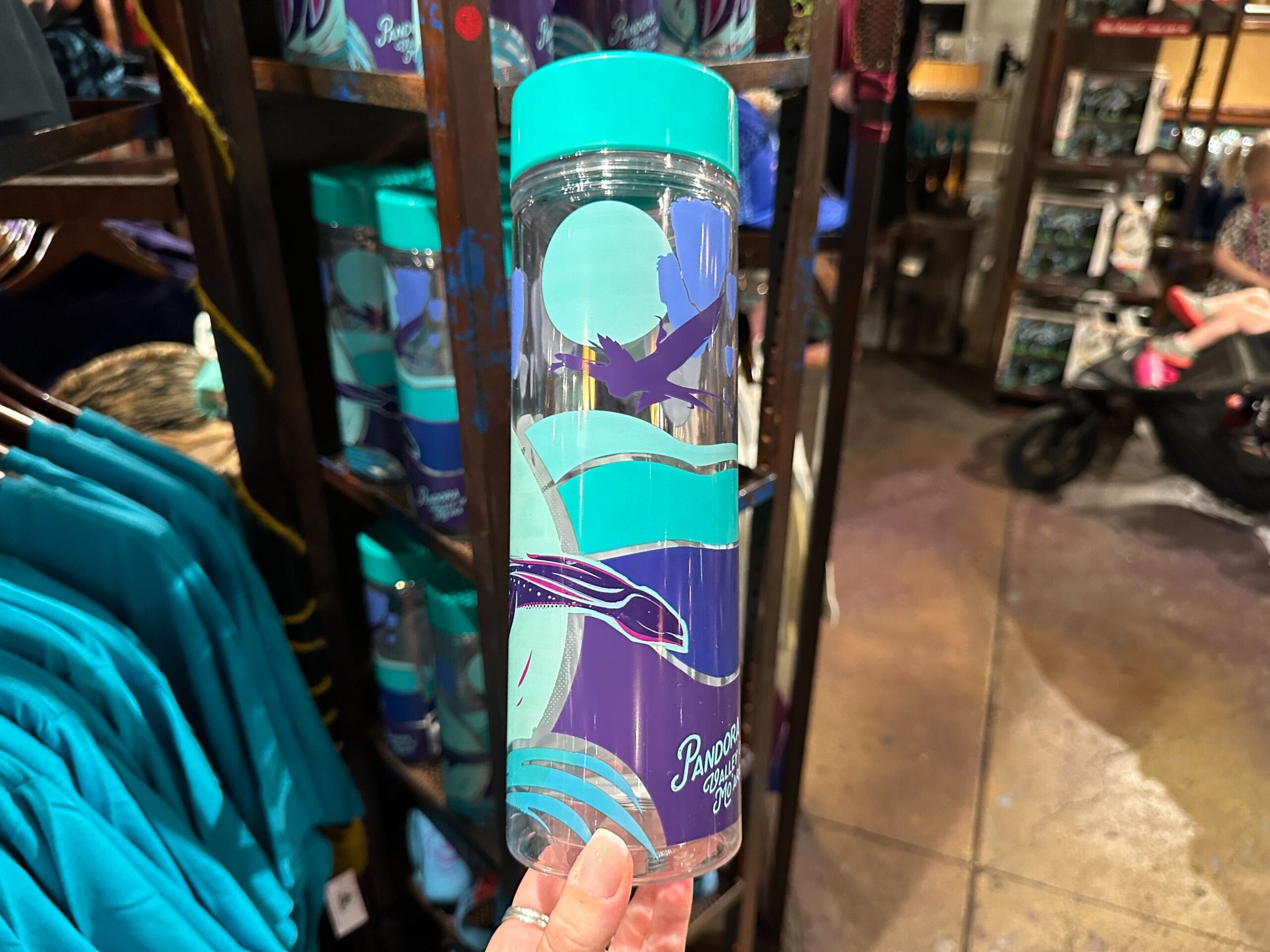 A hand holds a turquoise and purple Pandora merchandise water bottle with ocean and wave designs in a store.