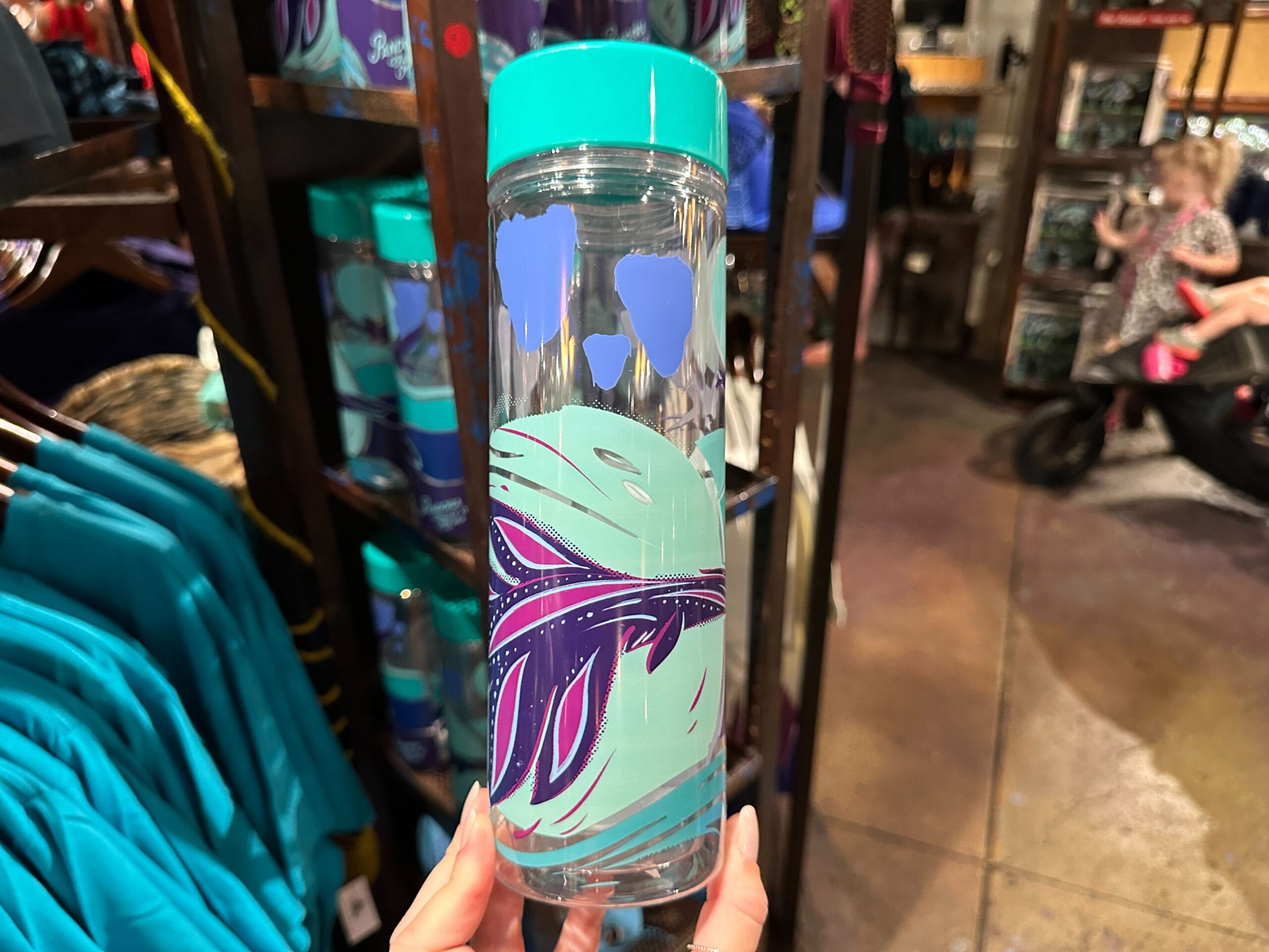 A hand holds a clear water bottle with teal and purple leaf designs, Pandora Merch style, and a teal lid in a store.