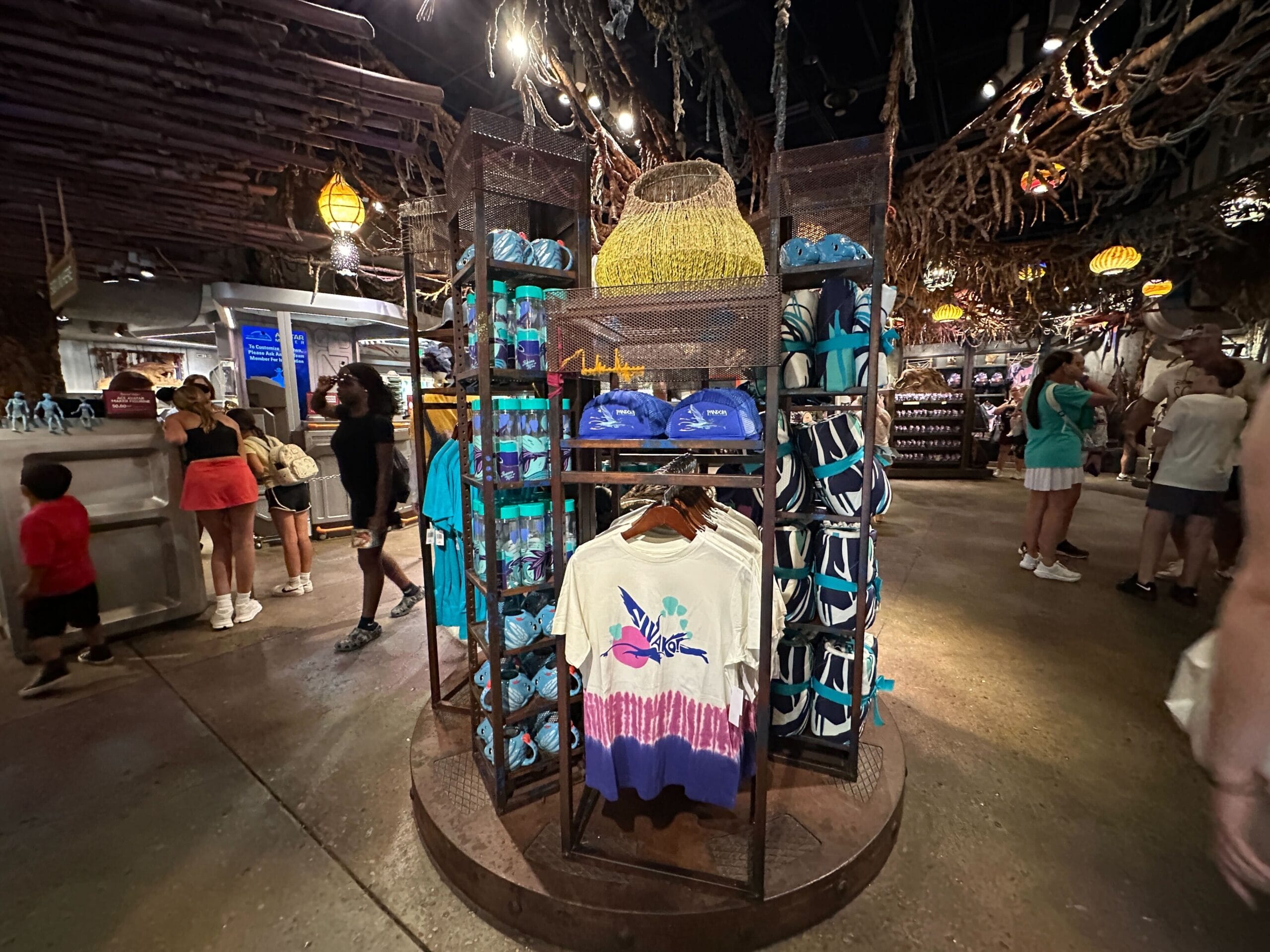 A store display of Pandora Merch—blue hats, white shirts, blue souvenirs—surrounded by shoppers in a dimly lit themed shop.