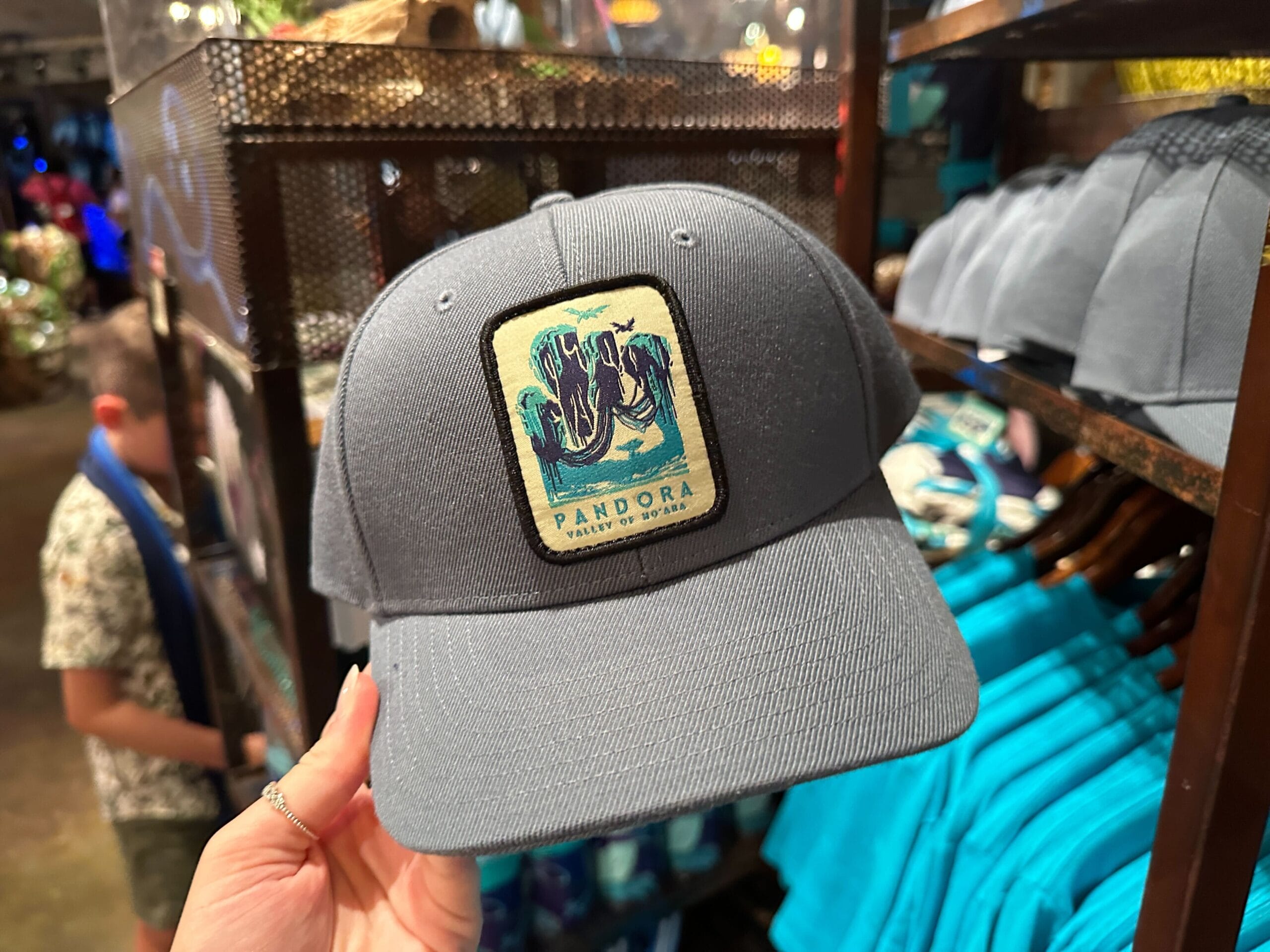 A hand holds a gray Pandora Merch cap with an illustrated patch in a store.