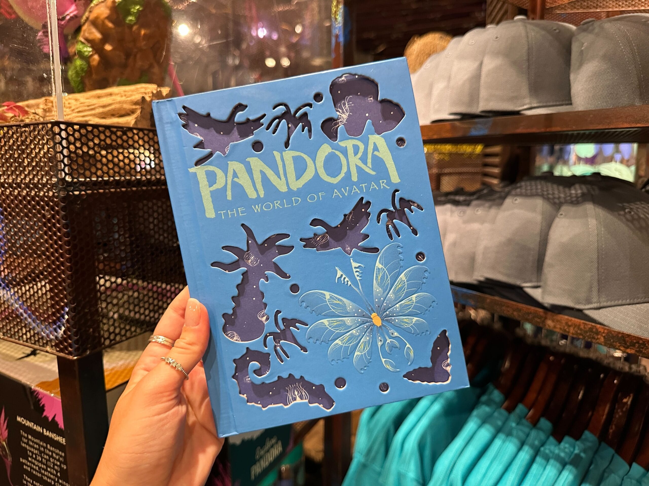 A hand holds a blue Pandora Merch journal with cutout designs inside a "Pandora: The World of Avatar" gift shop.