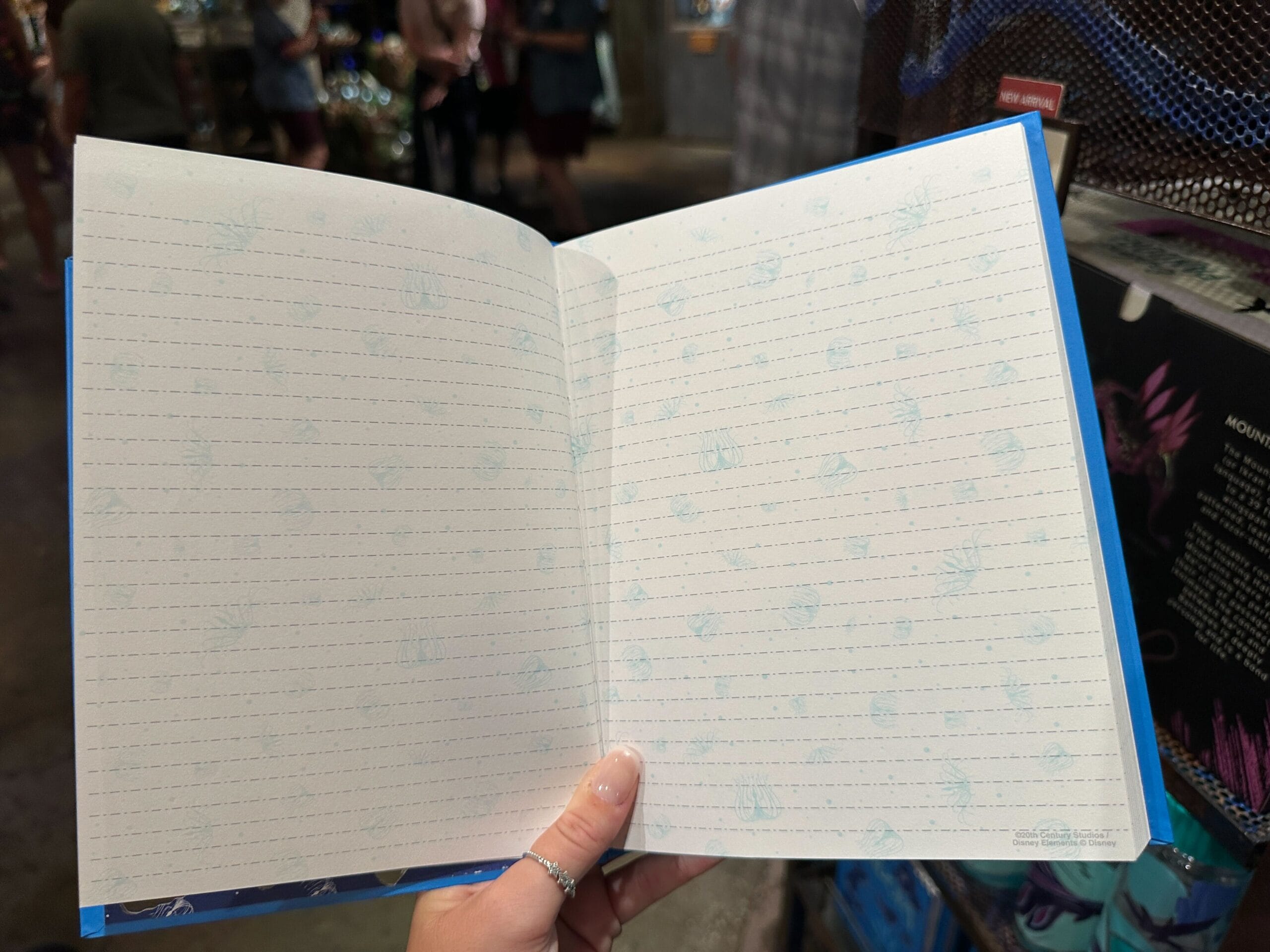 A hand holds open a lined notebook with faint blue patterns, perfect for jotting ideas or Pandora Merch notes in a store.