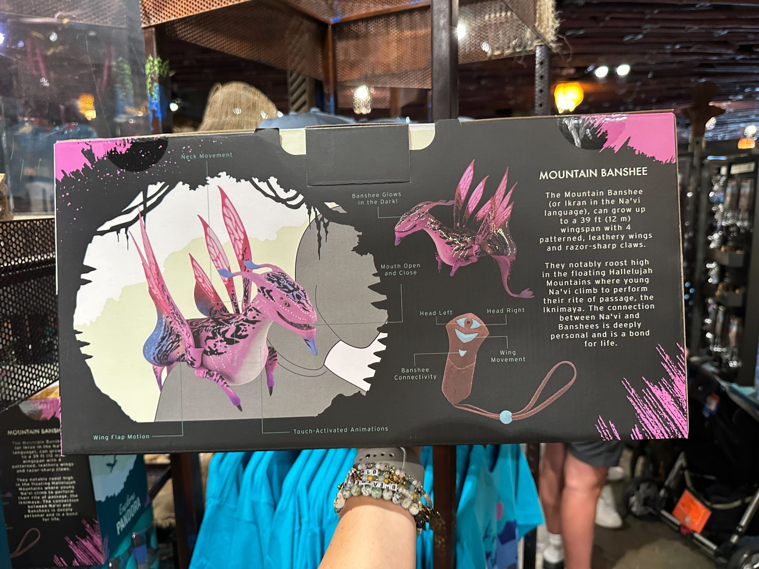 A hand holds a box displaying Pandora Merch—a toy Mountain Banshee—inside a shop filled with various items.