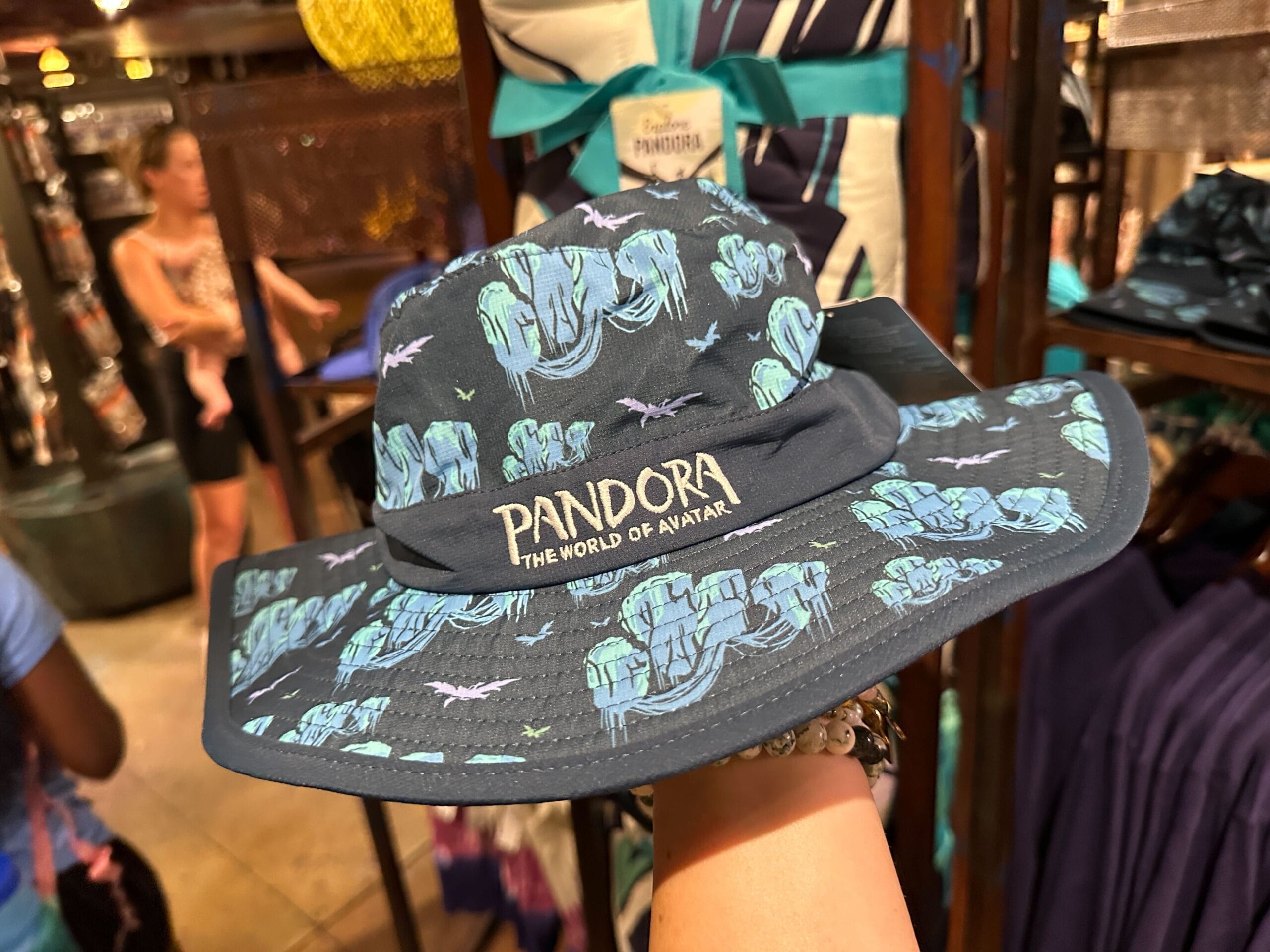 A hand holds Pandora Merch—a "Pandora: The World of Avatar" hat with blue pattern—in a merchandise store.
