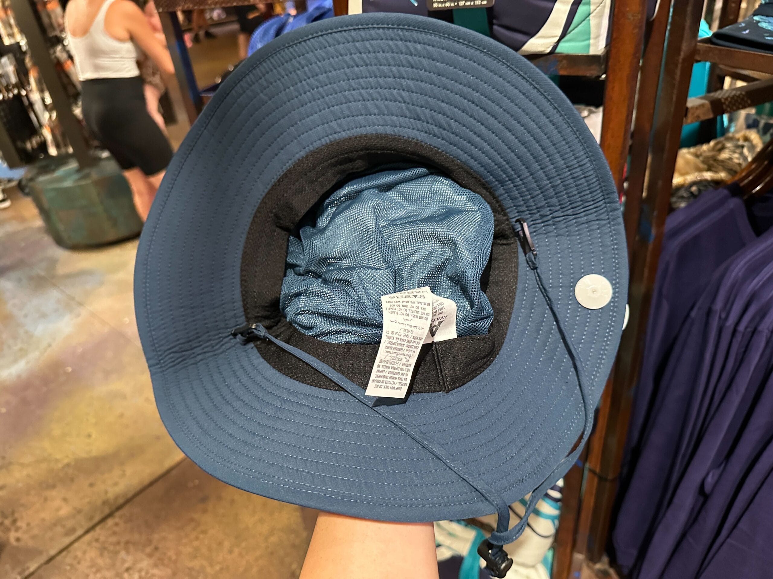 A hand holds a blue wide-brimmed Pandora Merch hat, showing its inside tags and chin strap in a clothing store.