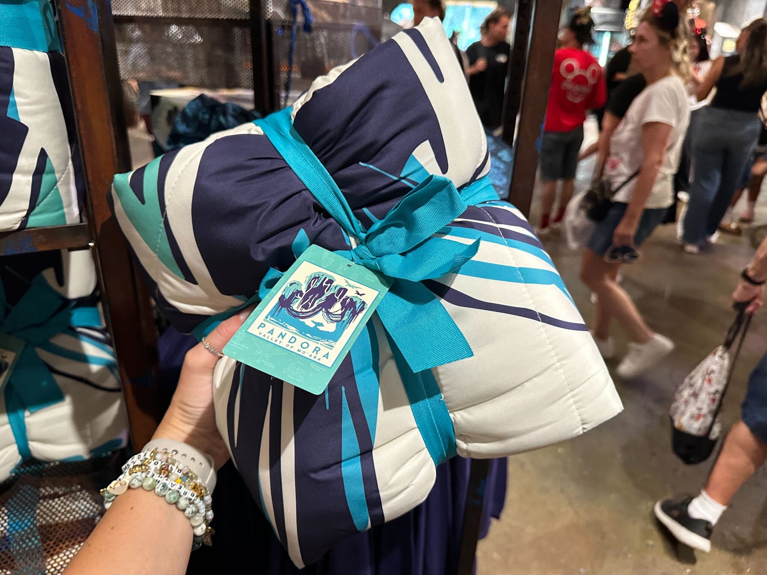 A hand holds a blue and white cushion with a turquoise ribbon and Pandora Merch tag in a busy store.