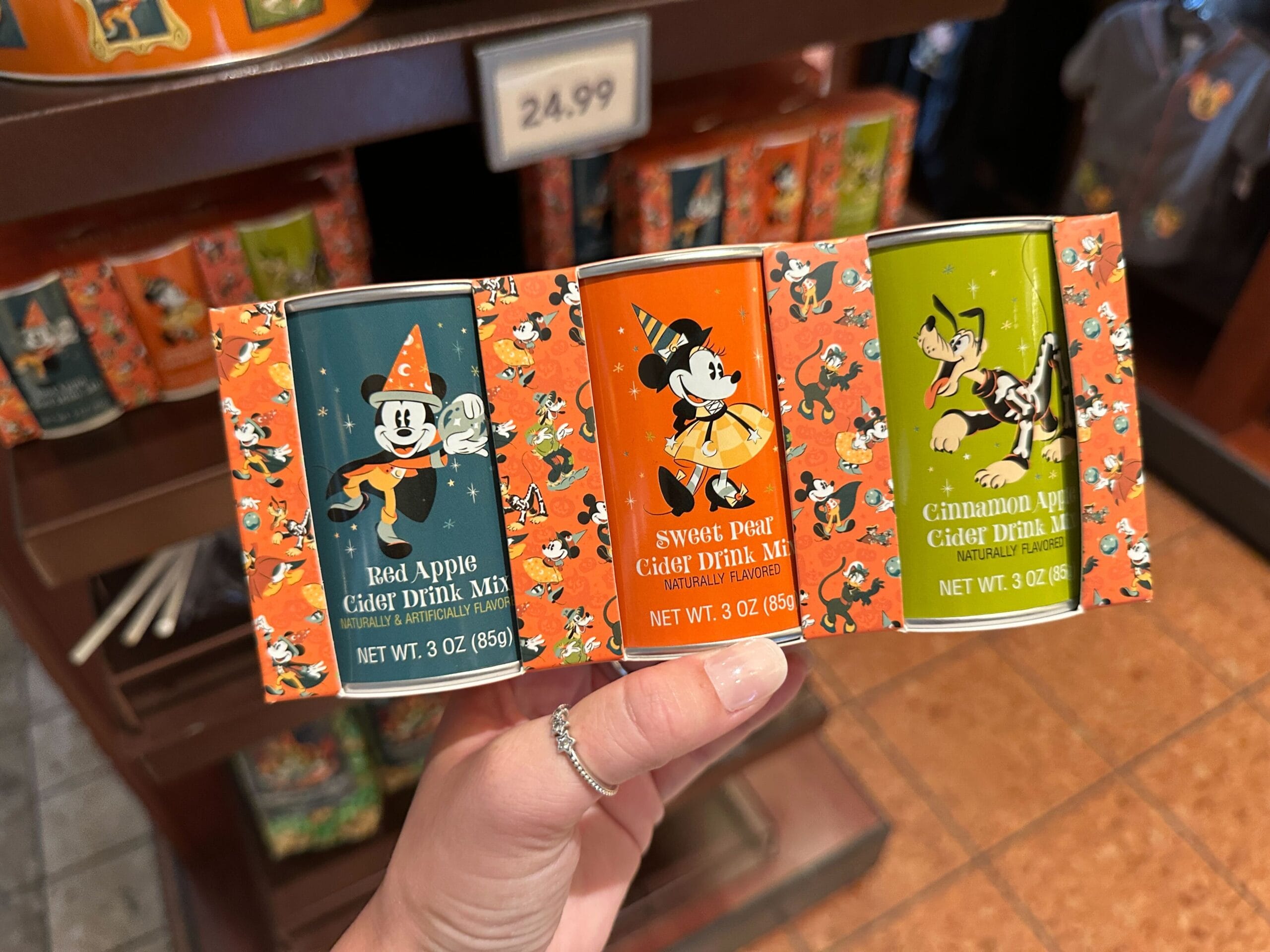 A hand holds three Disney Halloween Merch cider drink mixes with characters, priced at $24.99.
