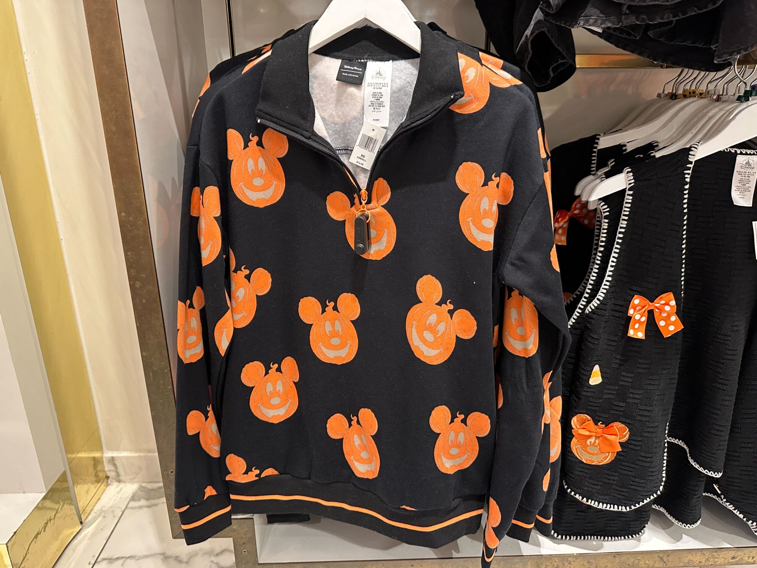 Black zip-up sweater with orange Mickey Mouse pumpkins hangs near a black Her Universe dress with orange accents.
