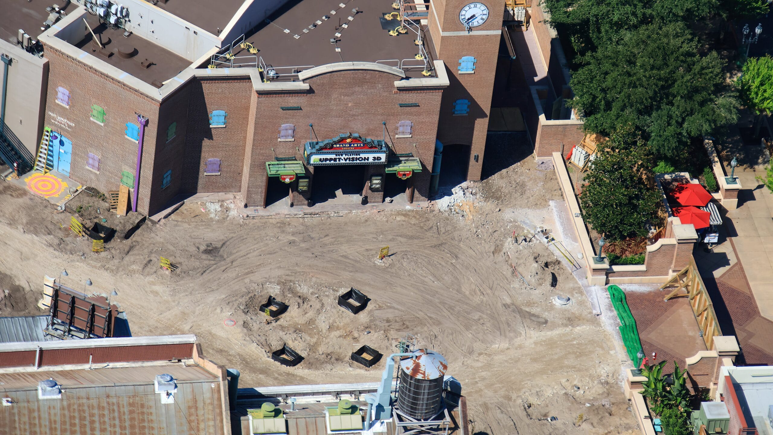 Aerial view of a construction site on flat dirt by a brick building with a clock and trees, resembling a Muppets scene.