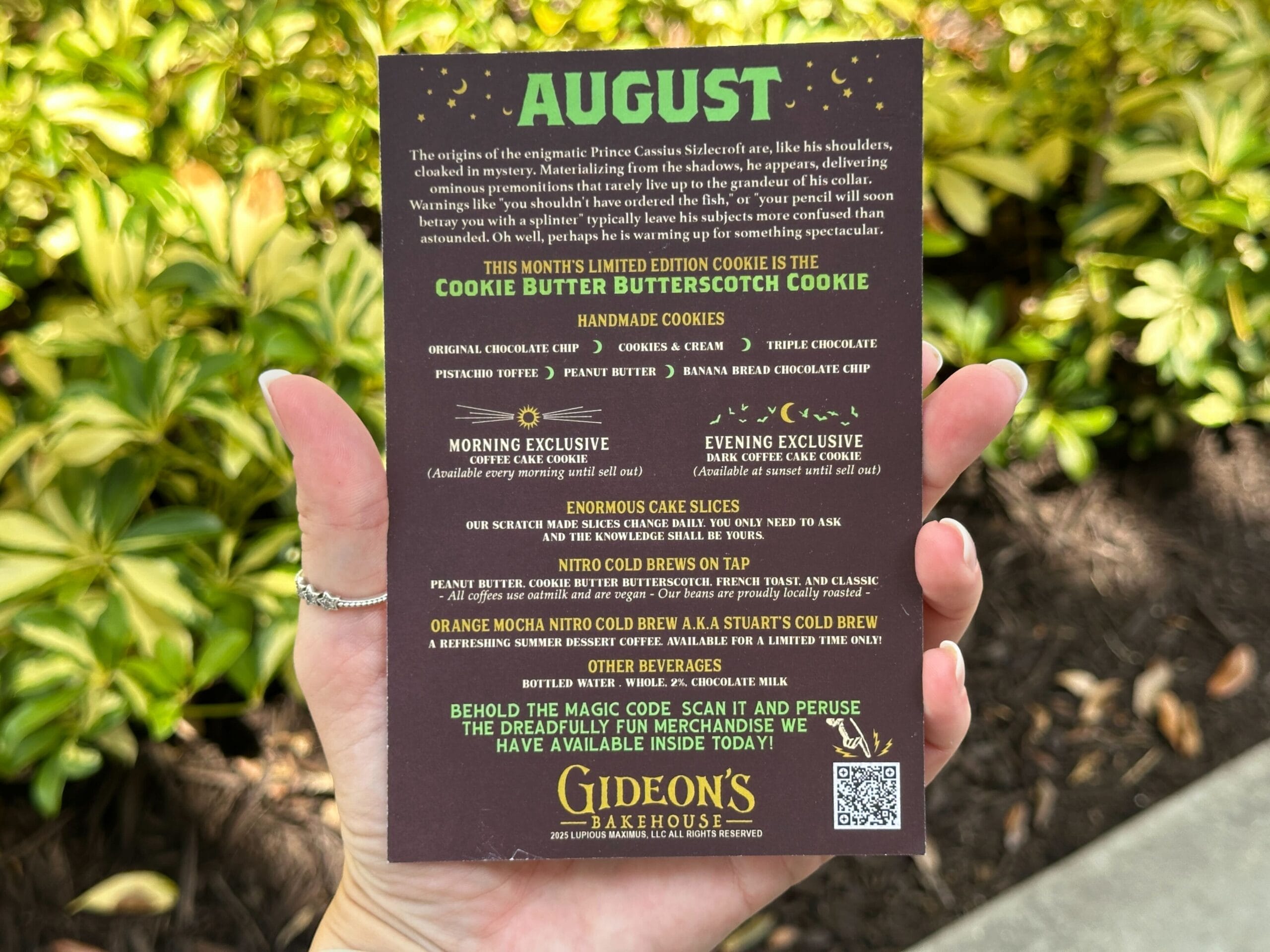 A hand holds an August menu from Gideon's Bakehouse, listing cookies and drinks, with greenery in the background.