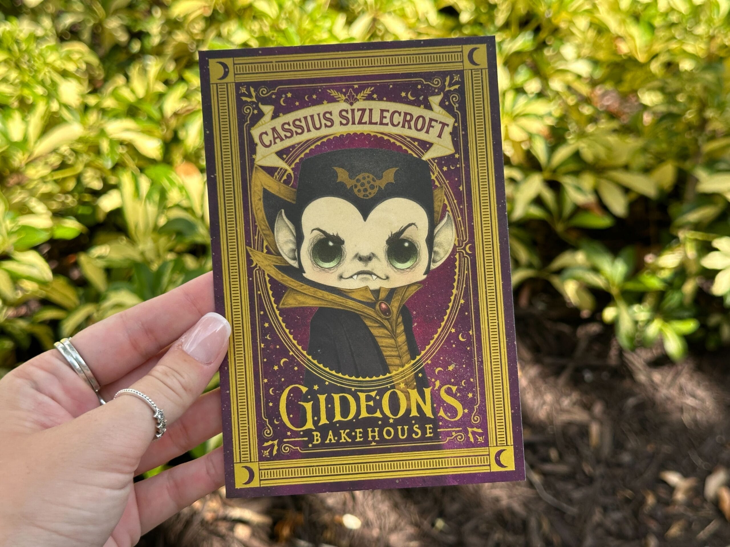 A hand holds a Gideon's Bakehouse card featuring a cartoon vampire named Cassius Sizlecroft, with plants in the background.