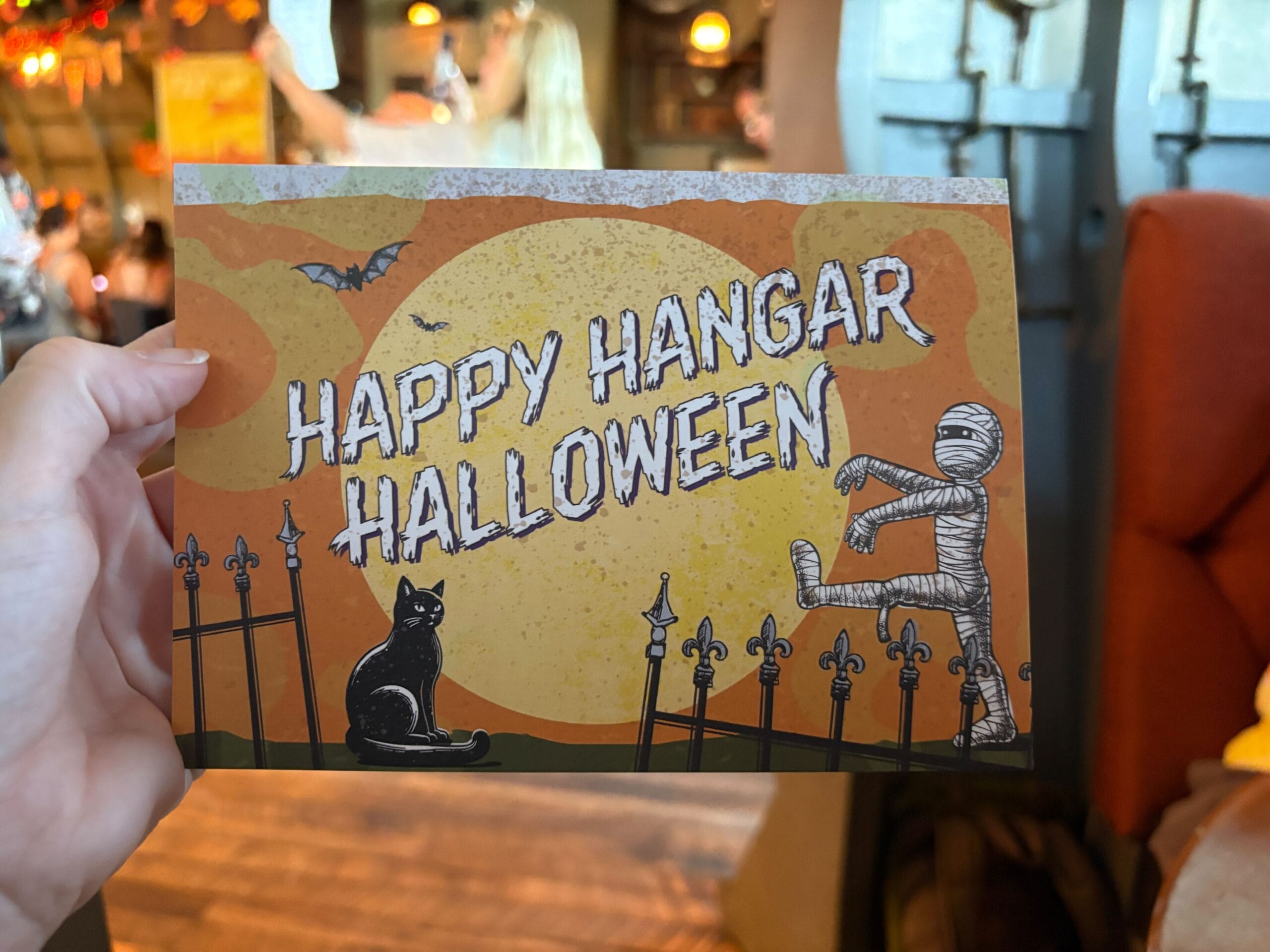 A hand holds a Halloween card from Jock Lindsey's Hangar Bar at Disney Springs with a black cat, mummy, bats, and a fence.